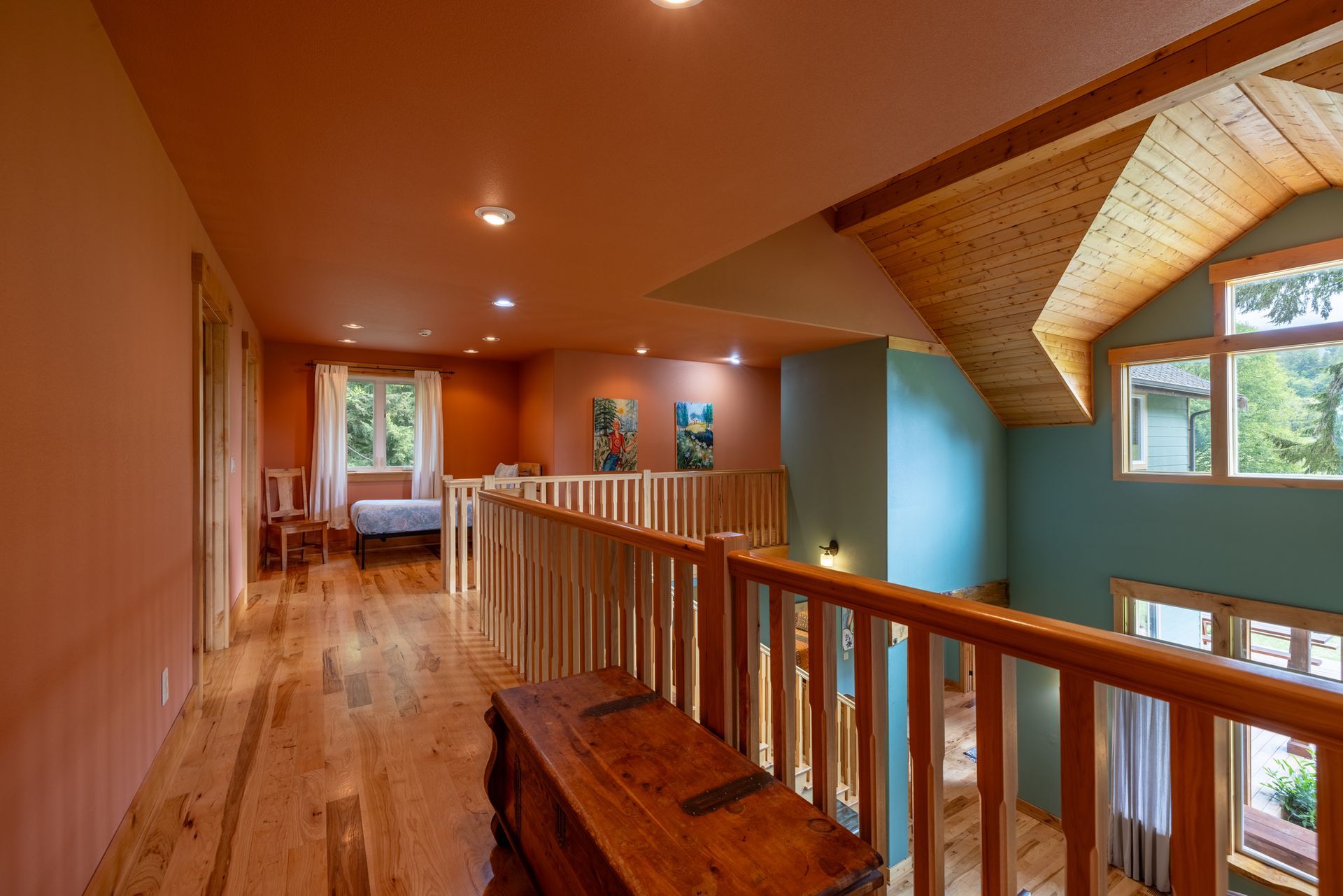 A wooden hallway with orange walls and a wood-beamed ceiling, leading to a balcony overlooking a teal-colored wall with windows.