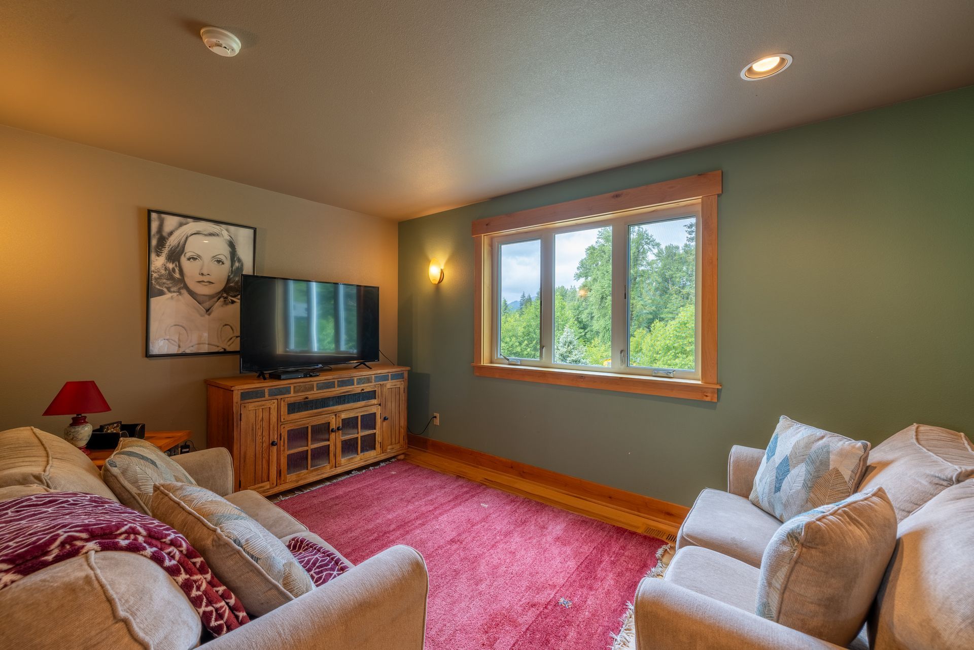 A cozy living room with a TV, two couches, a rug, and a window with a view of trees. The walls are green and tan.