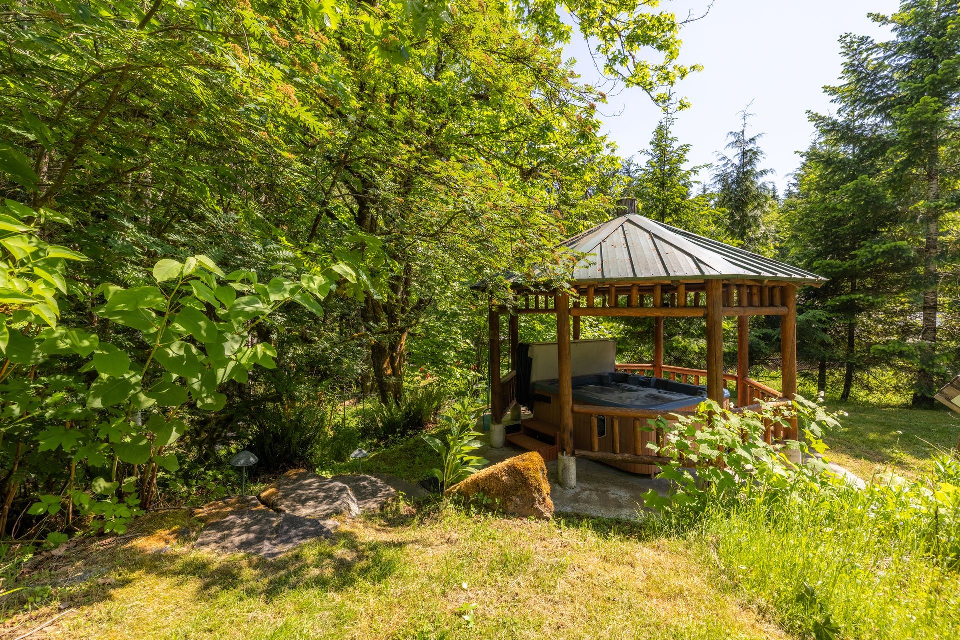 Wooden gazebo with a hot tub nestled in a lush, green forest setting. Sunny day with trees and vegetation surrounding it.