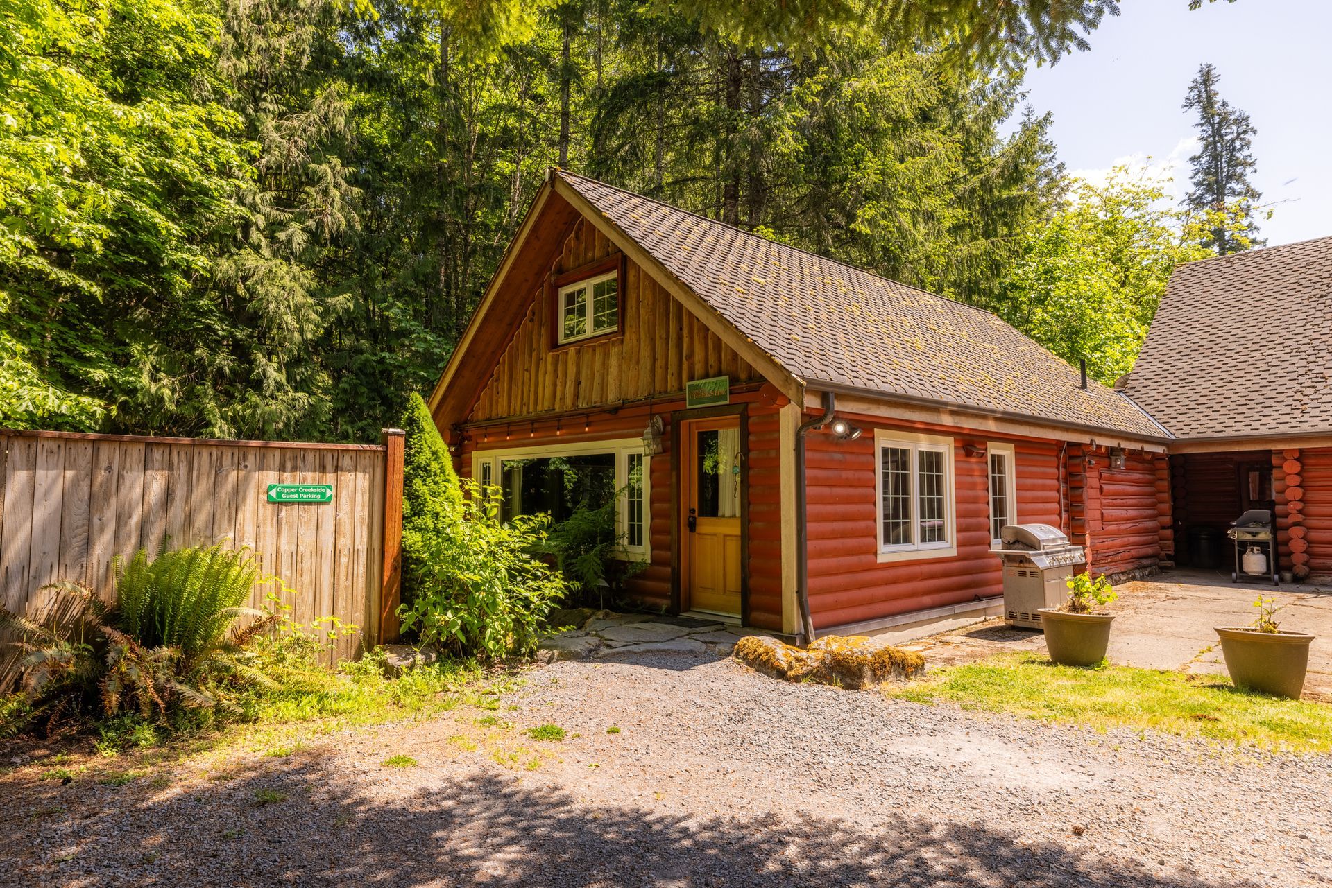 Rustic, red cabin nestled in a wooded area. The cabin has a gravel driveway, wooden fence, and a grill.