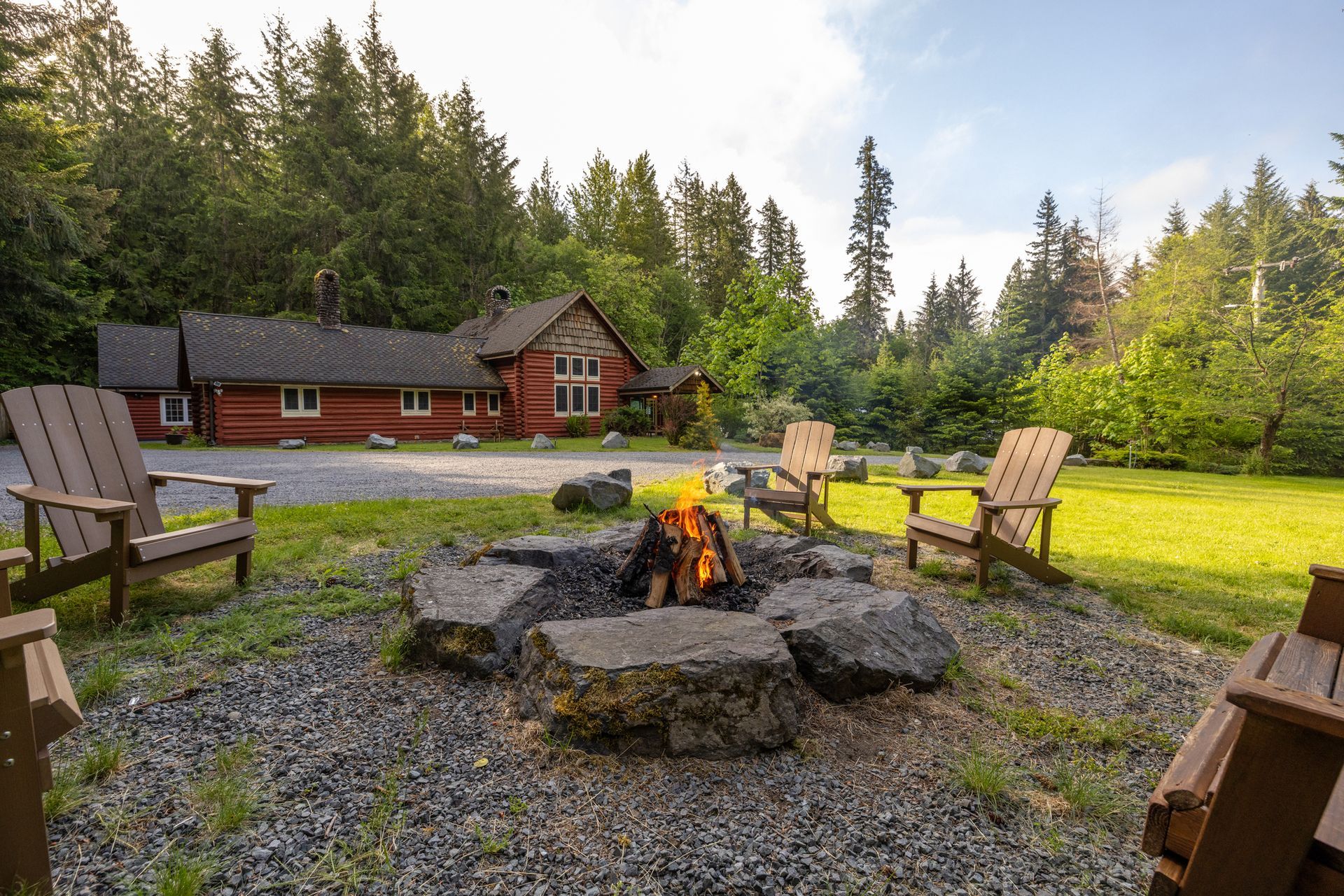 A fire pit with burning flames surrounded by wooden chairs on a gravel area in front of a red cabin in a forest.