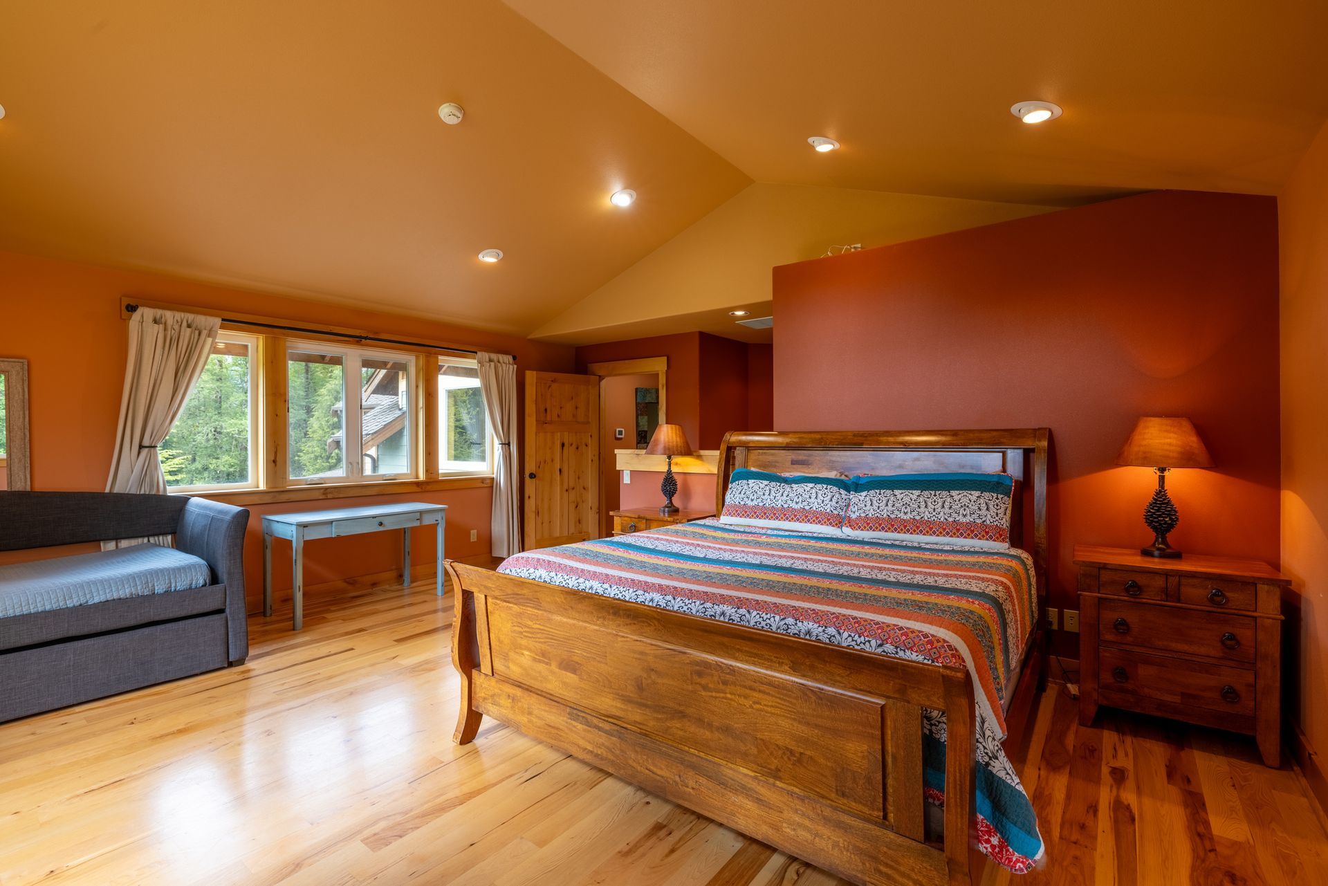 Bright bedroom with wooden furniture and hardwood floors. Includes a bed, dresser, sofa, and desk near a window. The walls are orange.