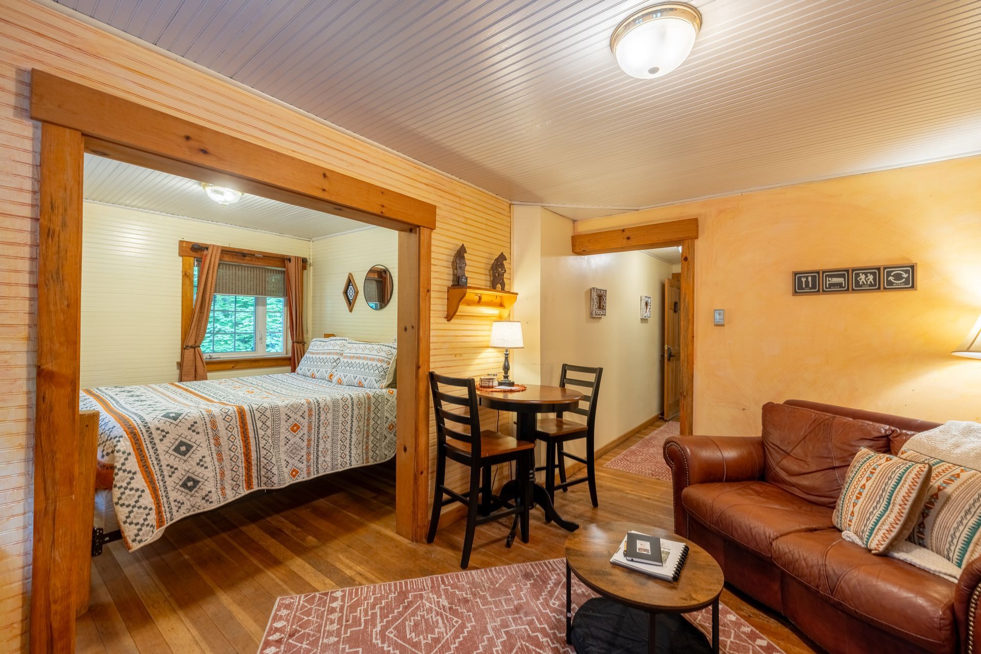 Cozy cabin interior: bed in a room, living area with sofa, dining table, and an entryway. Wooden floors, warm tones.