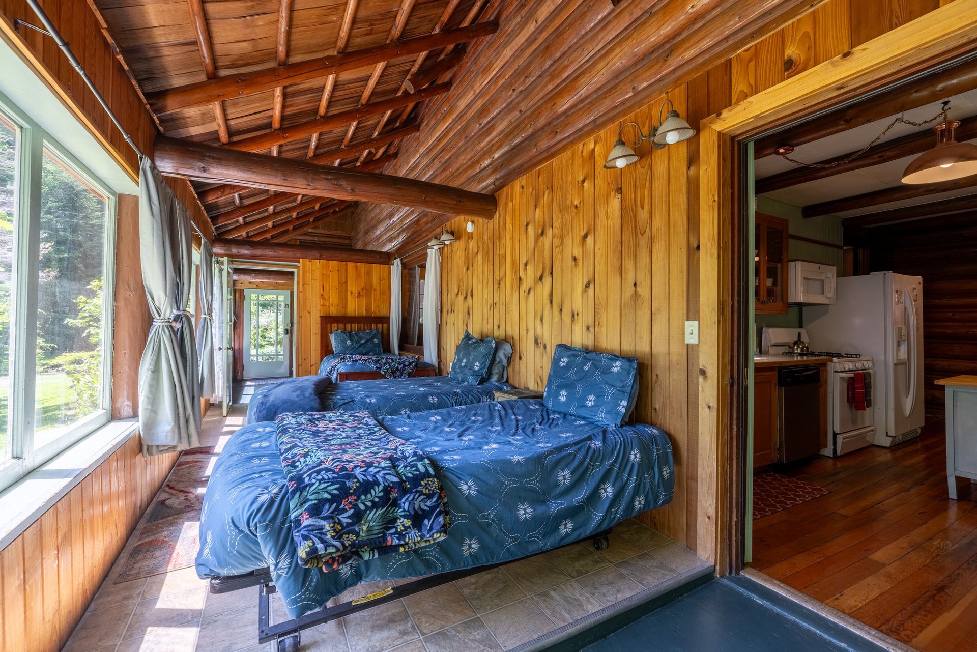Sunroom with two twin beds, wooden walls and ceiling, and a sliding door leading to a kitchen.