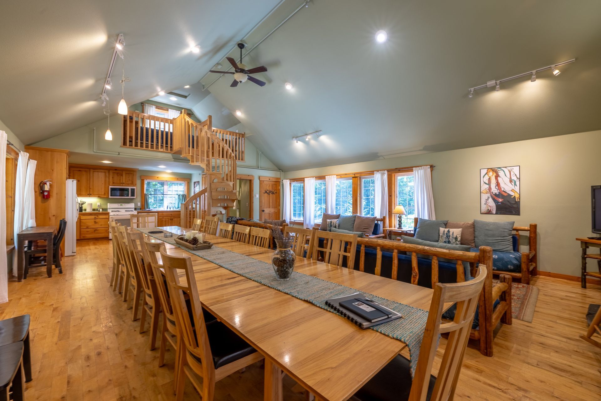 Spacious cabin interior with a long dining table and multiple seating areas. Pine flooring, high ceilings, and a loft are visible.