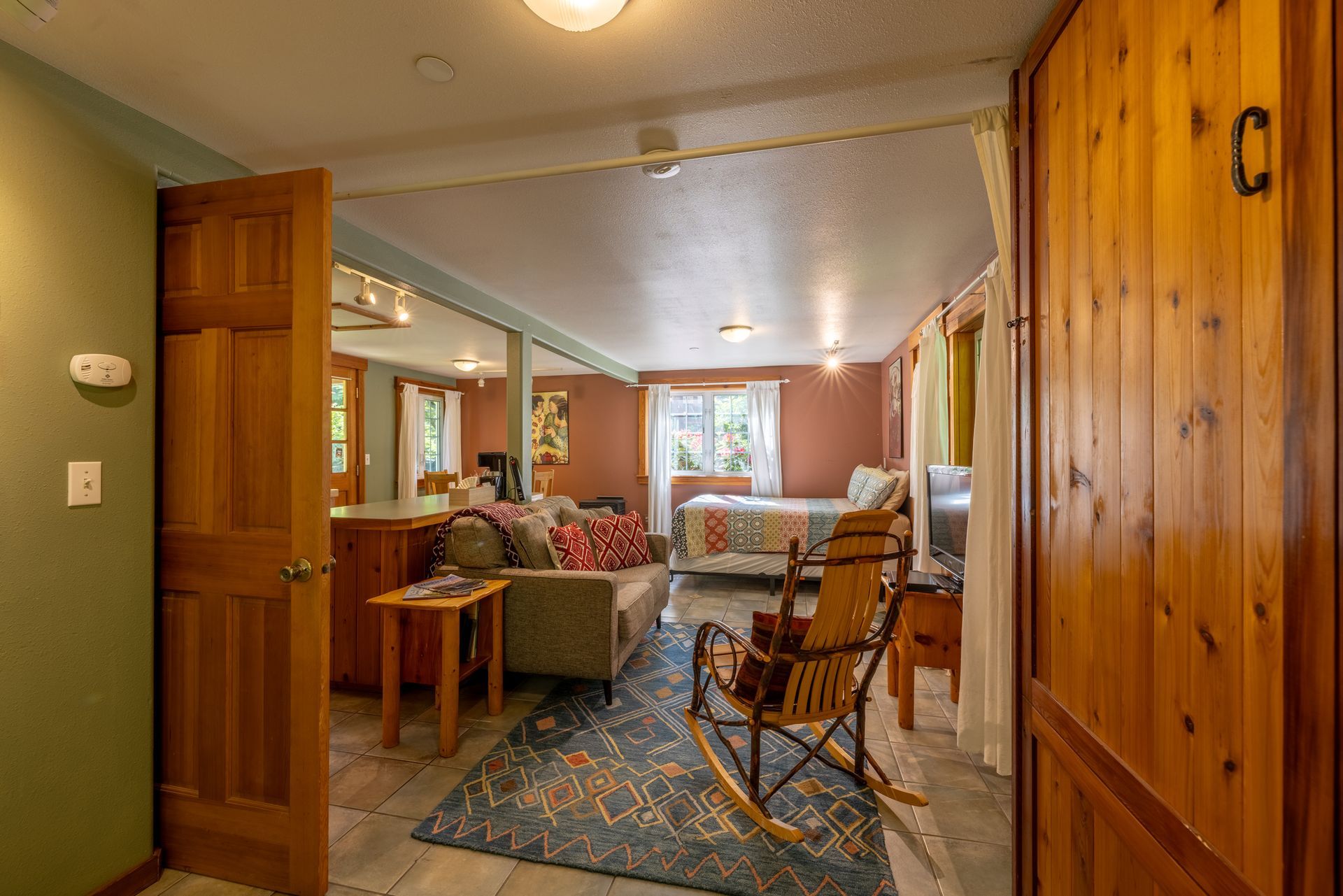 A doorway leads into a rustic room with a couch, bed, and rocking chair. Soft natural light illuminates the space.