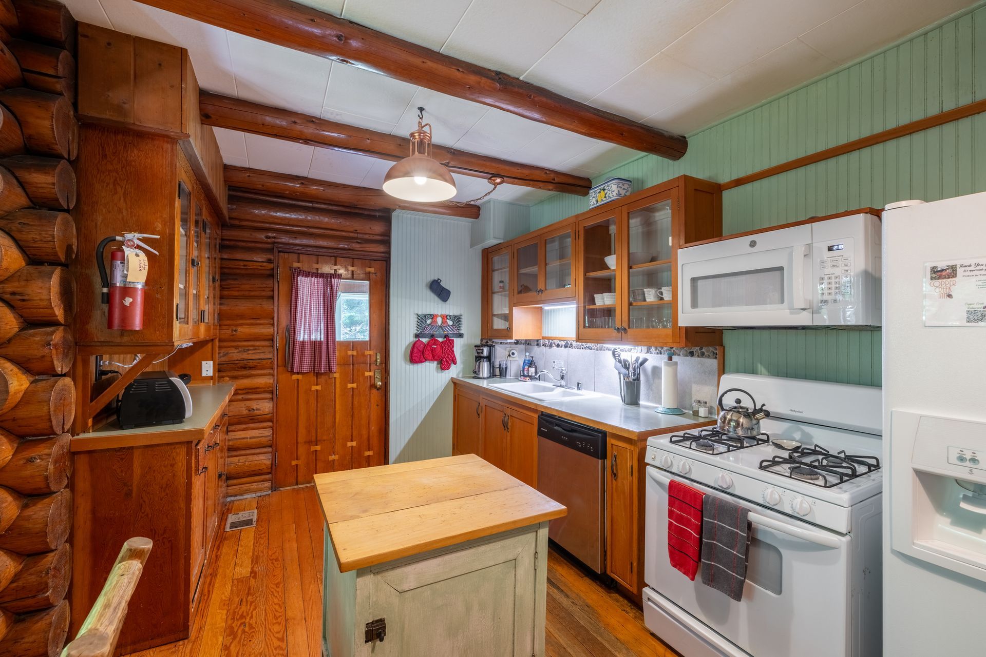 Rustic kitchen with wooden beams, cabinets, and a small island. Appliances include a white range, microwave, and refrigerator.