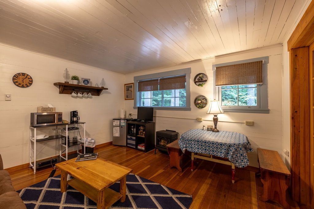 Cozy living space with wood floors, a blue rug, a wooden coffee table, and a small dining table near the windows.