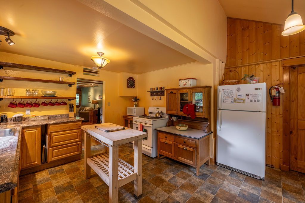 A warm-toned kitchen with wooden cabinets, countertops, and an island. A refrigerator, microwave, and open doorway are visible.