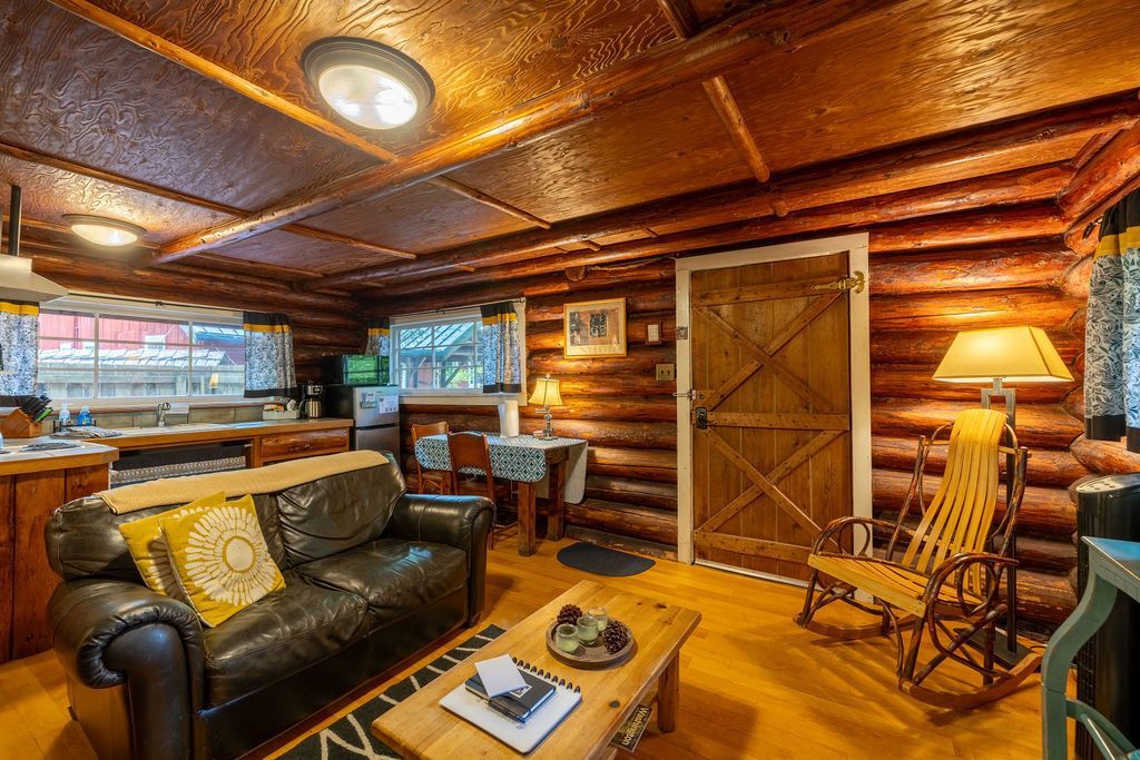 Cozy cabin interior with wood paneling, a leather sofa, rocking chair, and a wooden door. Warm lighting and rustic decor create a welcoming atmosphere.