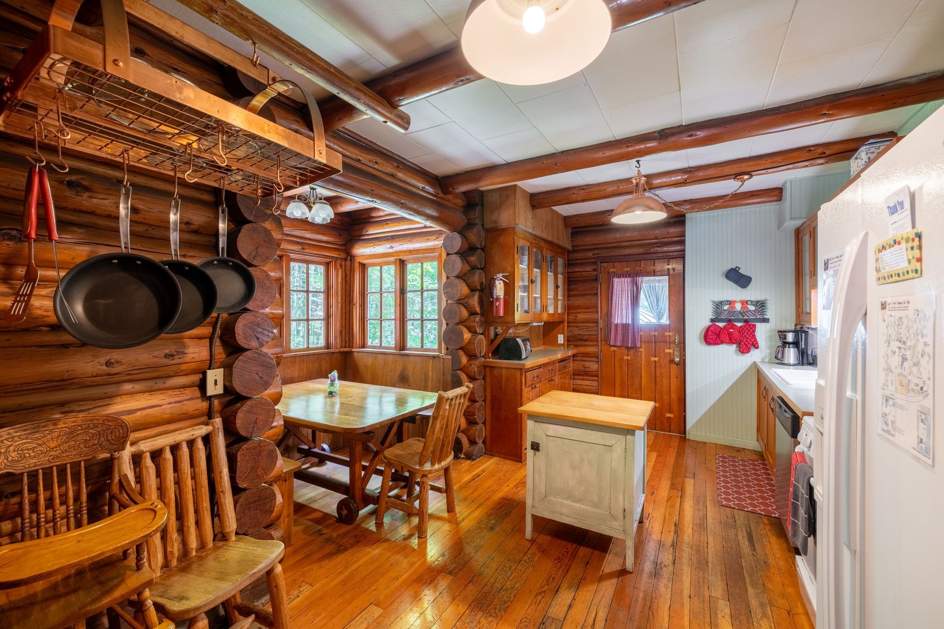 Rustic kitchen with log walls and wooden floor. Includes a table, stove, and hanging pots.
