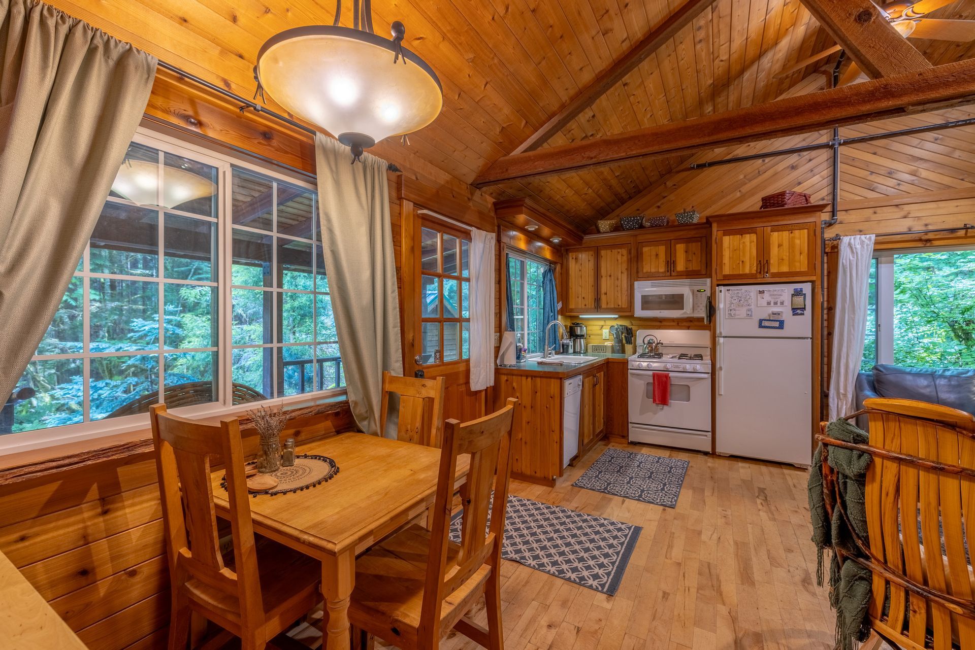 Cozy cabin interior with wood paneling, a dining table set for two, and a small kitchen with a window view of lush greenery.