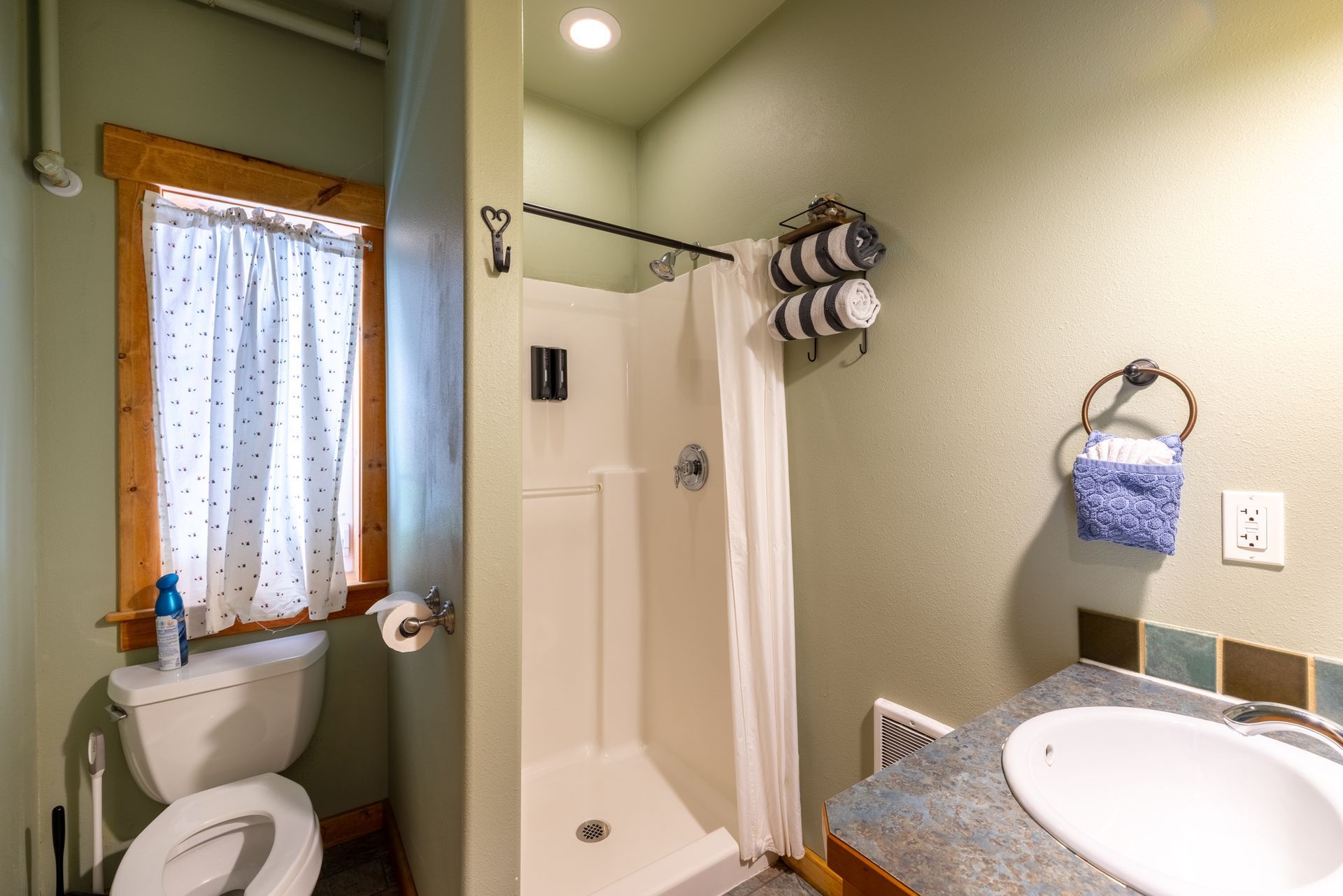 A small bathroom with a white shower stall, toilet, and sink. The walls are light green, and there's a small window with a curtain.