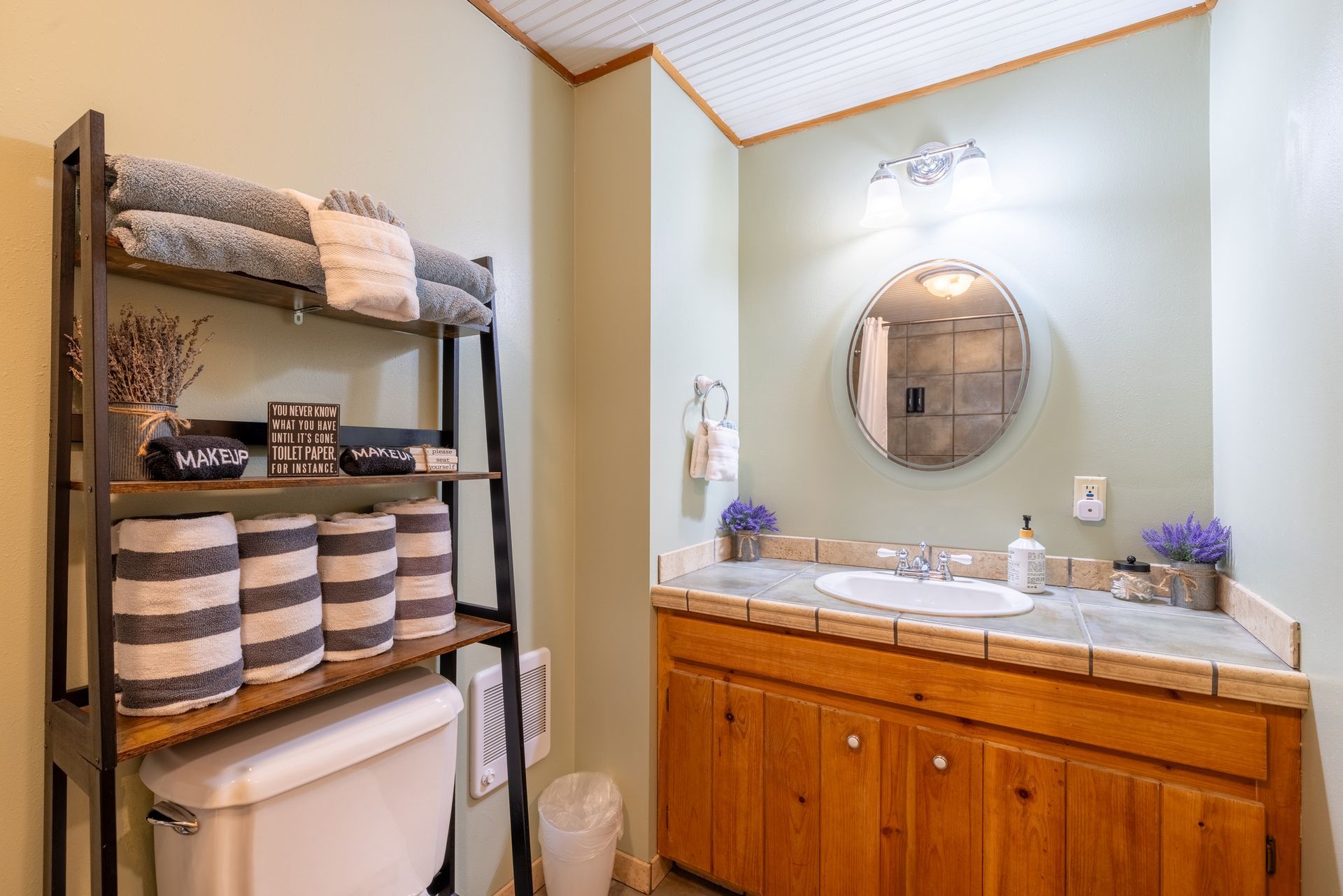 Small bathroom with a wooden vanity, round mirror, and a shelf holding towels and decorative items. Green walls and light-colored countertop.