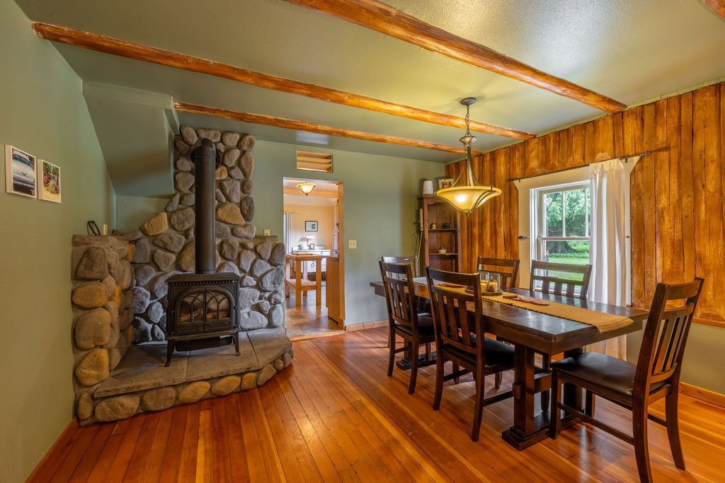 A rustic dining room with a wooden table and chairs, wood paneling, and a stone fireplace with a wood-burning stove.