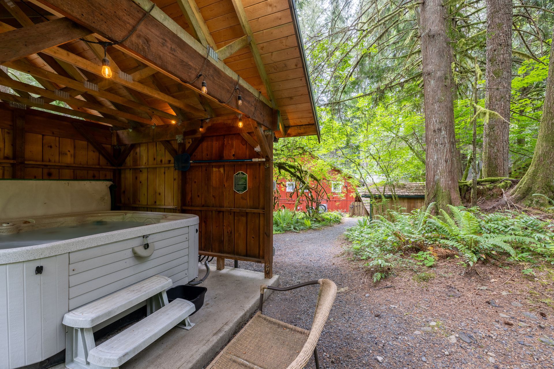 A hot tub under a wooden gazebo in a forest setting, with a path leading towards more buildings.