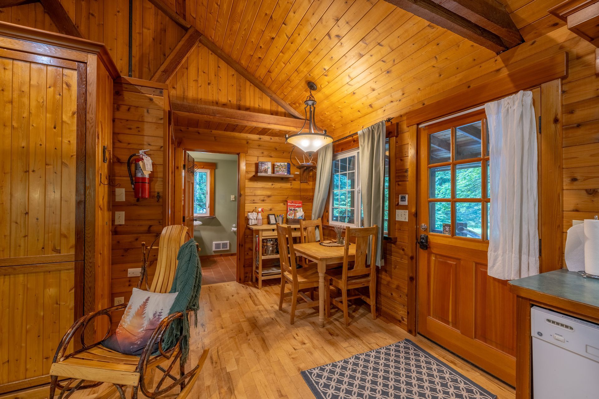 Interior of a log cabin with wooden walls, flooring, and vaulted ceiling. Dining area with a table, chairs, and a view into another room.