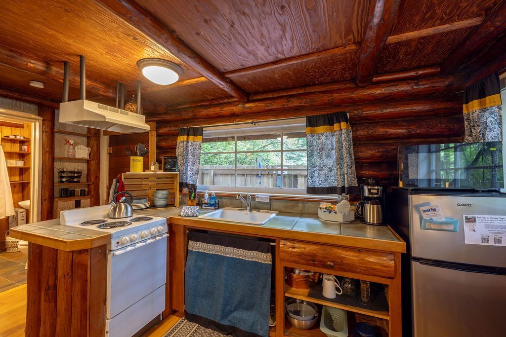 Log cabin kitchen with wooden beams, appliances, and a window. Features a stove, sink, refrigerator, and open shelving.