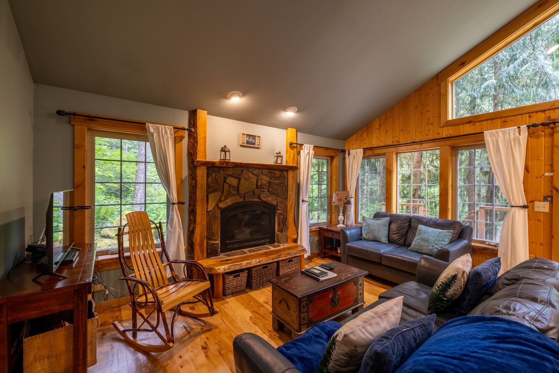 Cozy cabin living room with a stone fireplace, wooden walls, a gray sofa, and large windows overlooking trees.