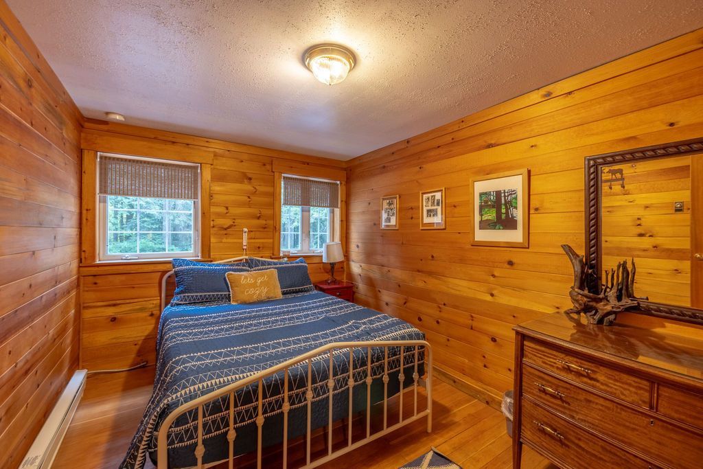 A cozy bedroom with wood-paneled walls, a bed with blue bedding, two windows, and a wooden dresser with a mirror.