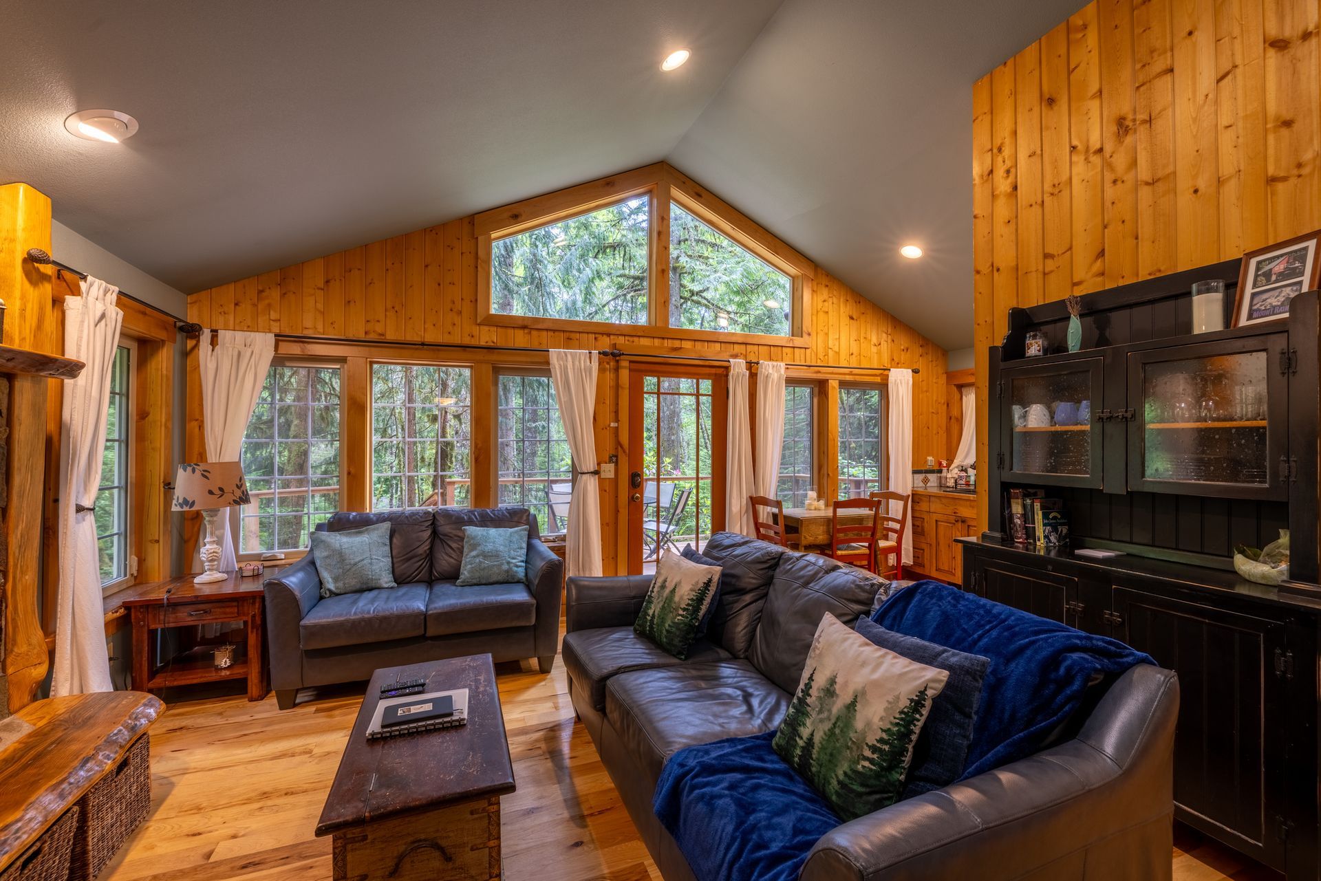 Living room with wood paneling, large windows, and two couches. Sunlight streams in, illuminating the space and furniture.