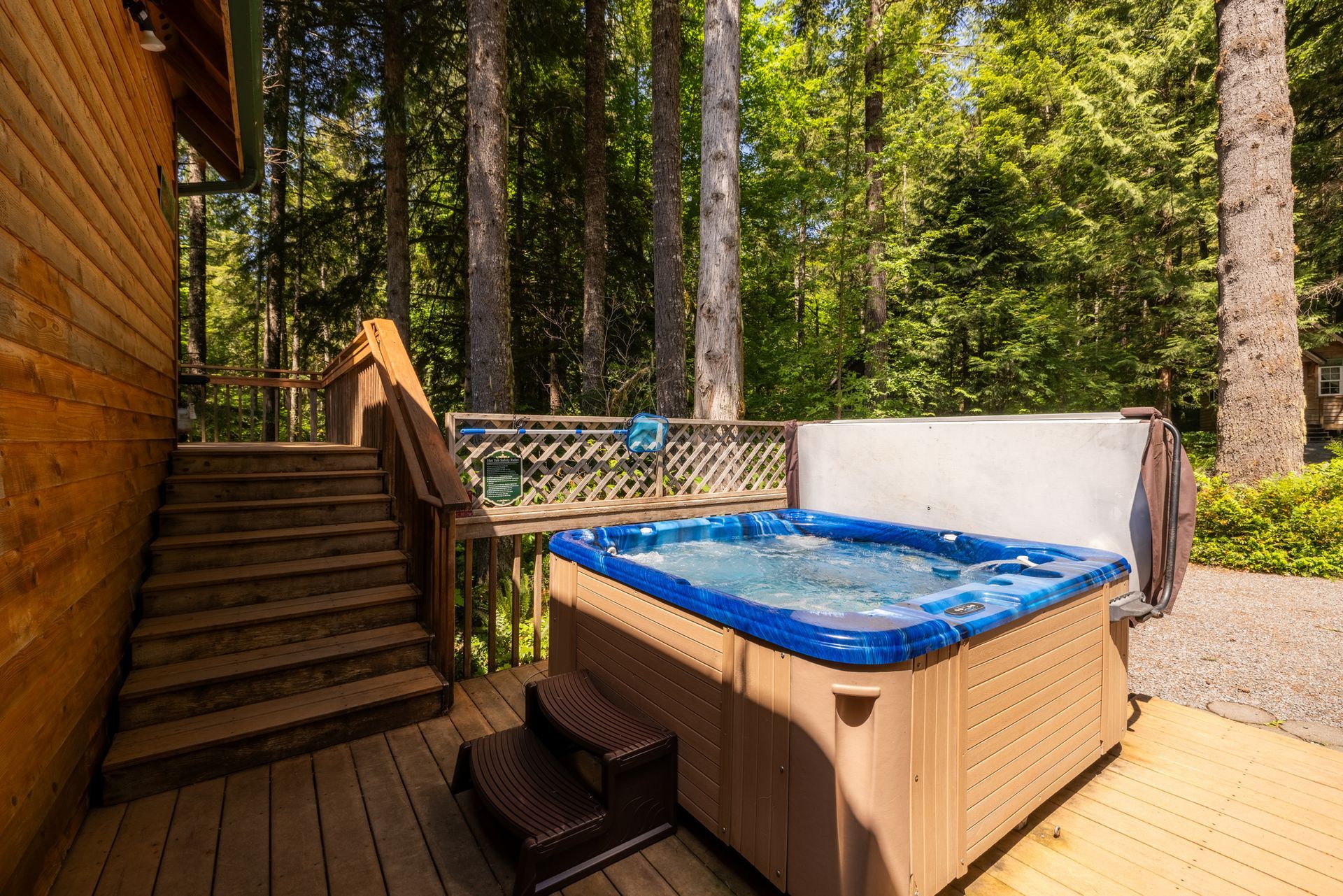 Hot tub on a wooden deck next to a staircase, surrounded by trees. The tub is blue and tan, filled with bubbling water.