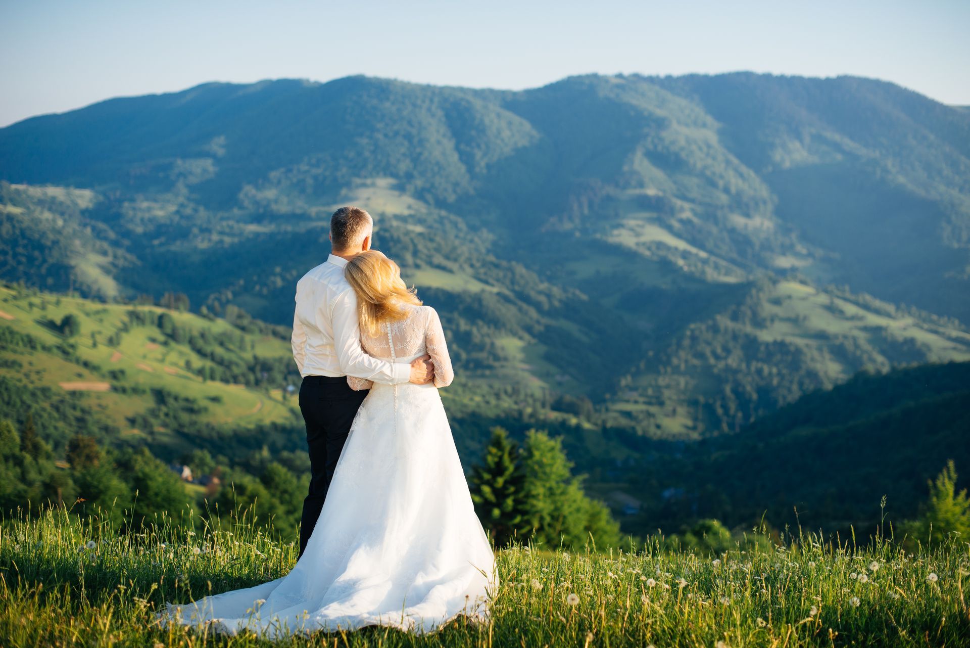 A bride and groom are standing in a field with mountains in the background.