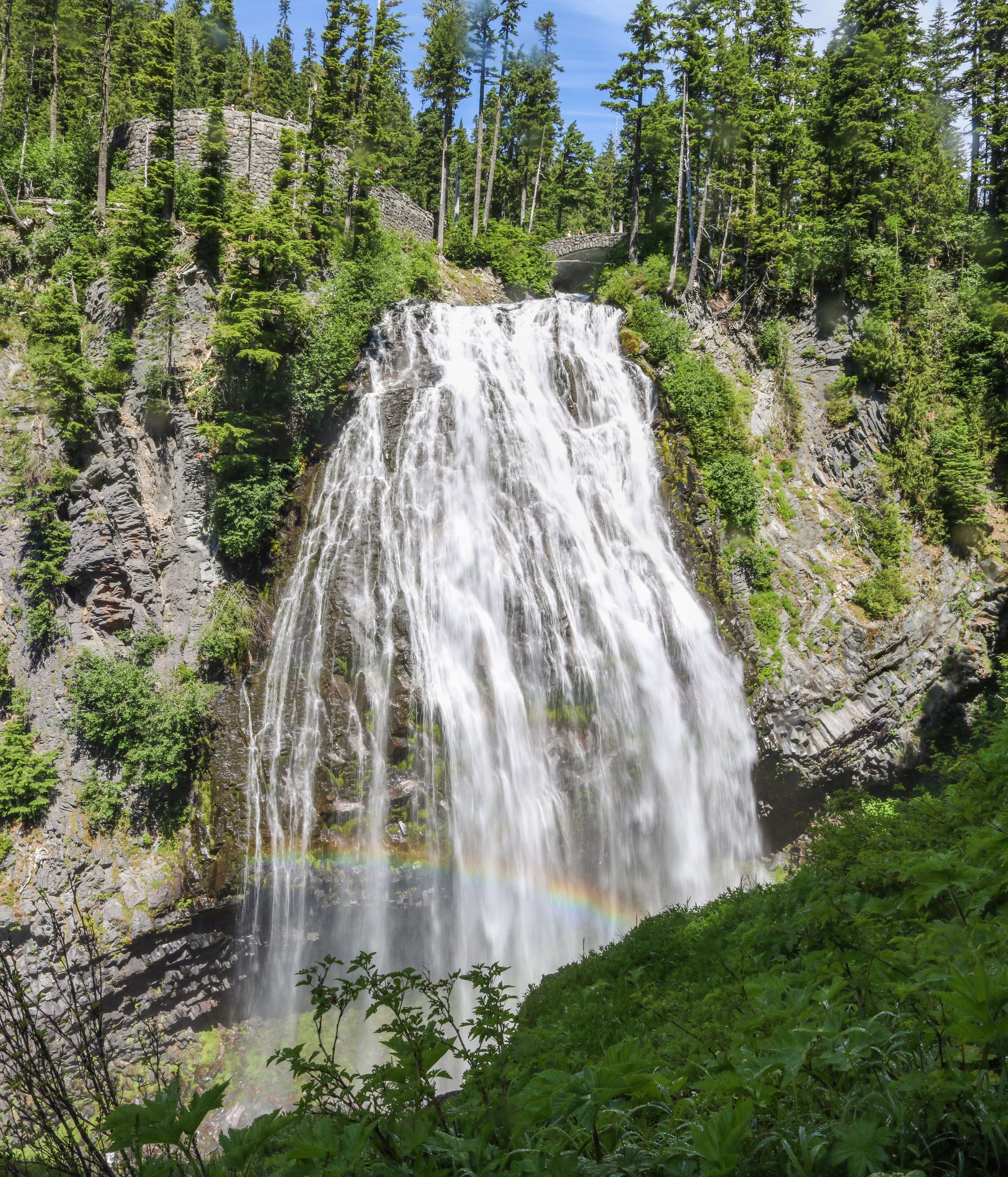 Waterfall cascading down rocky cliff, with a rainbow visible at the base. Surrounded by green trees.