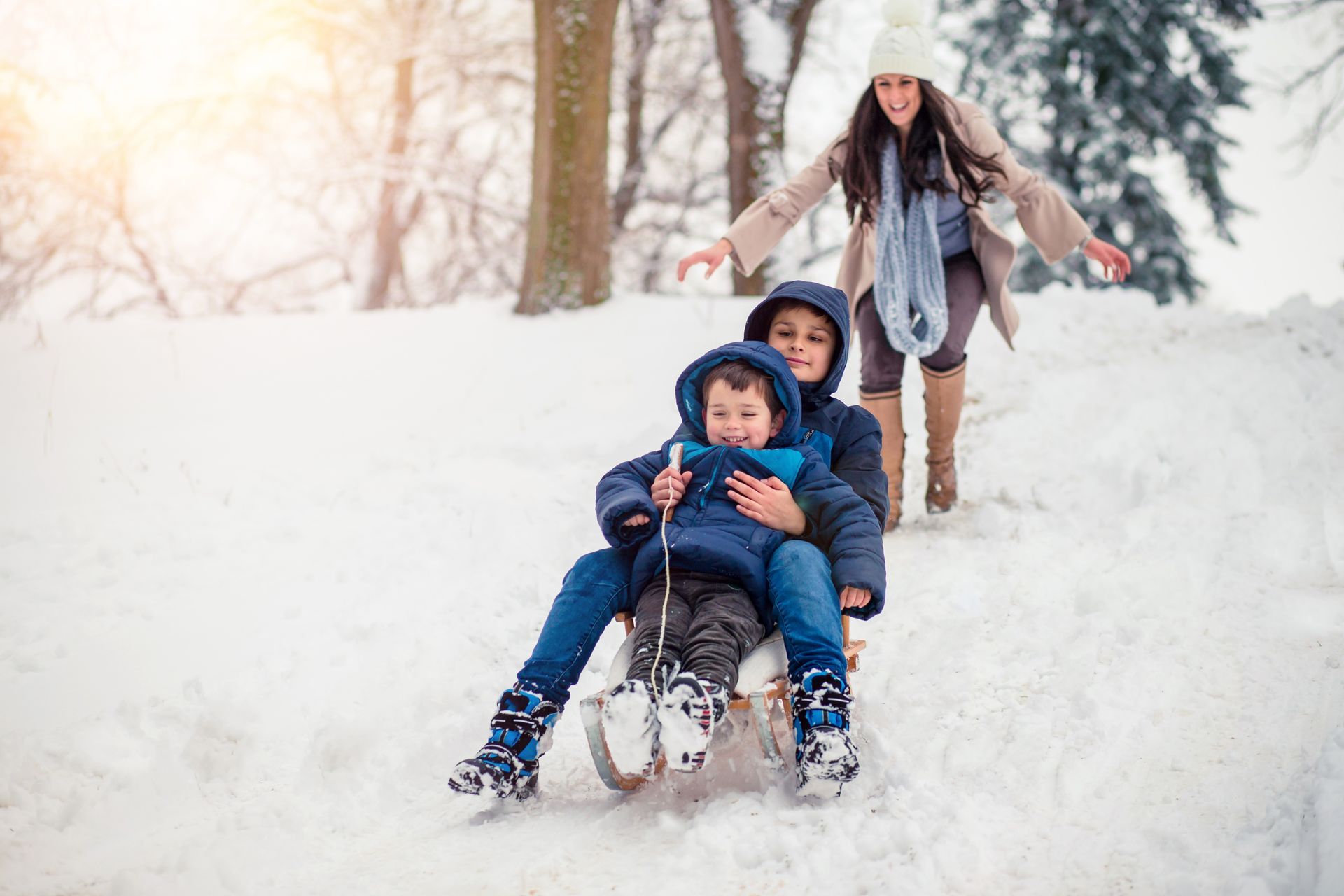 Woman pushing a sled down a snowy hill with a child; another woman trails behind, smiling.