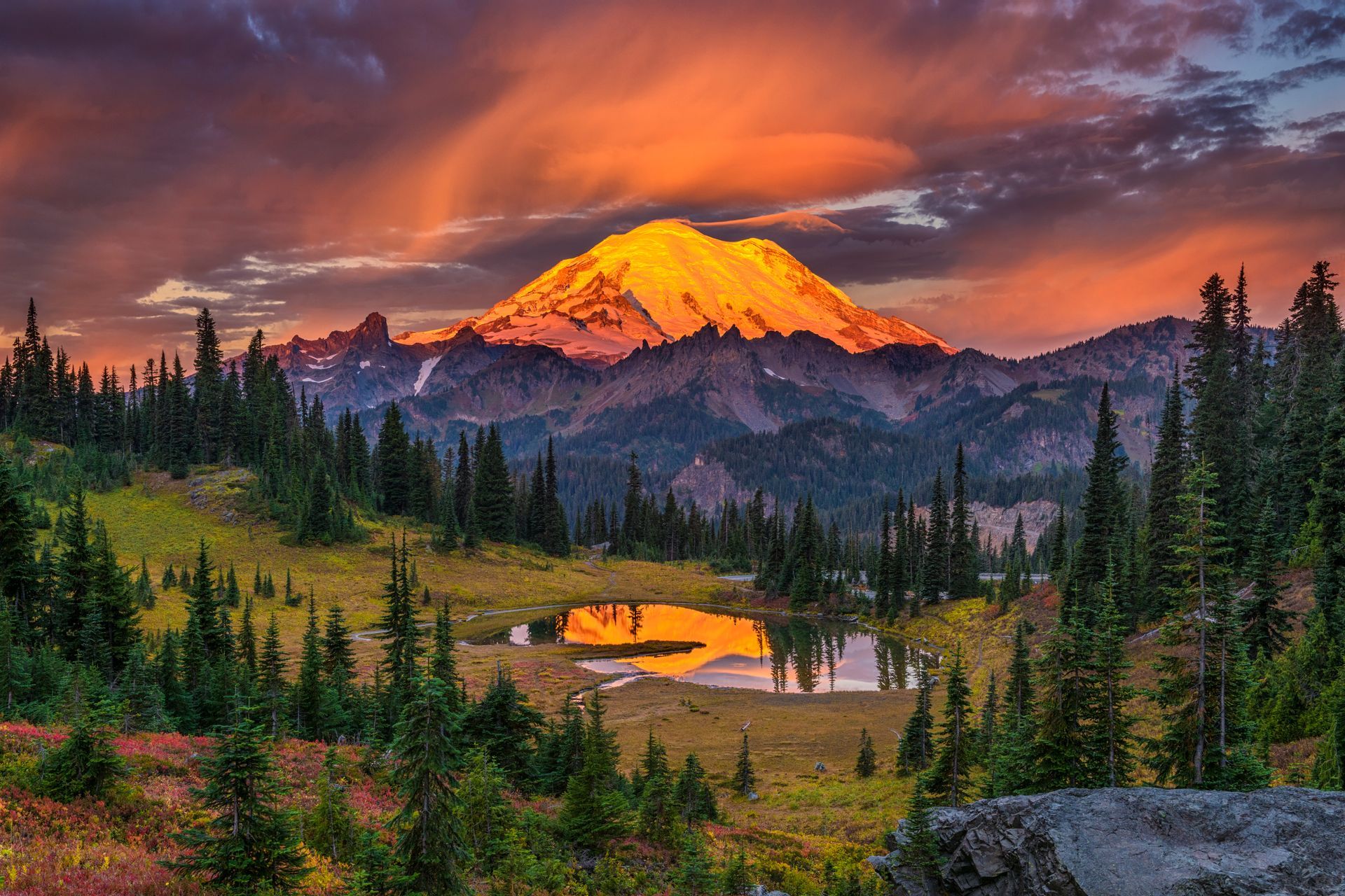 A mountain covered in snow is surrounded by trees and a lake at sunset.