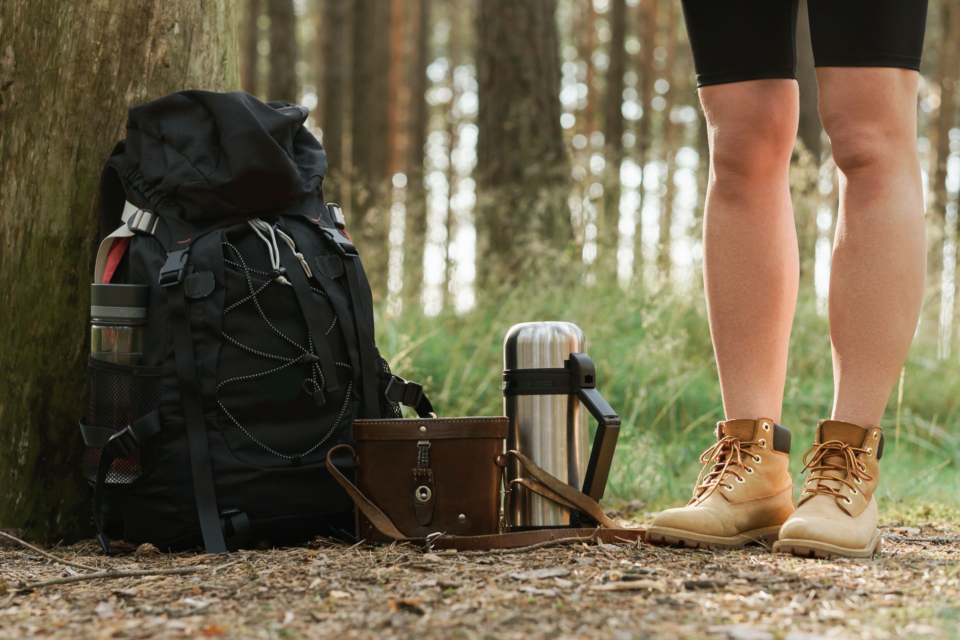 A person is standing next to a backpack and a thermos in the woods.