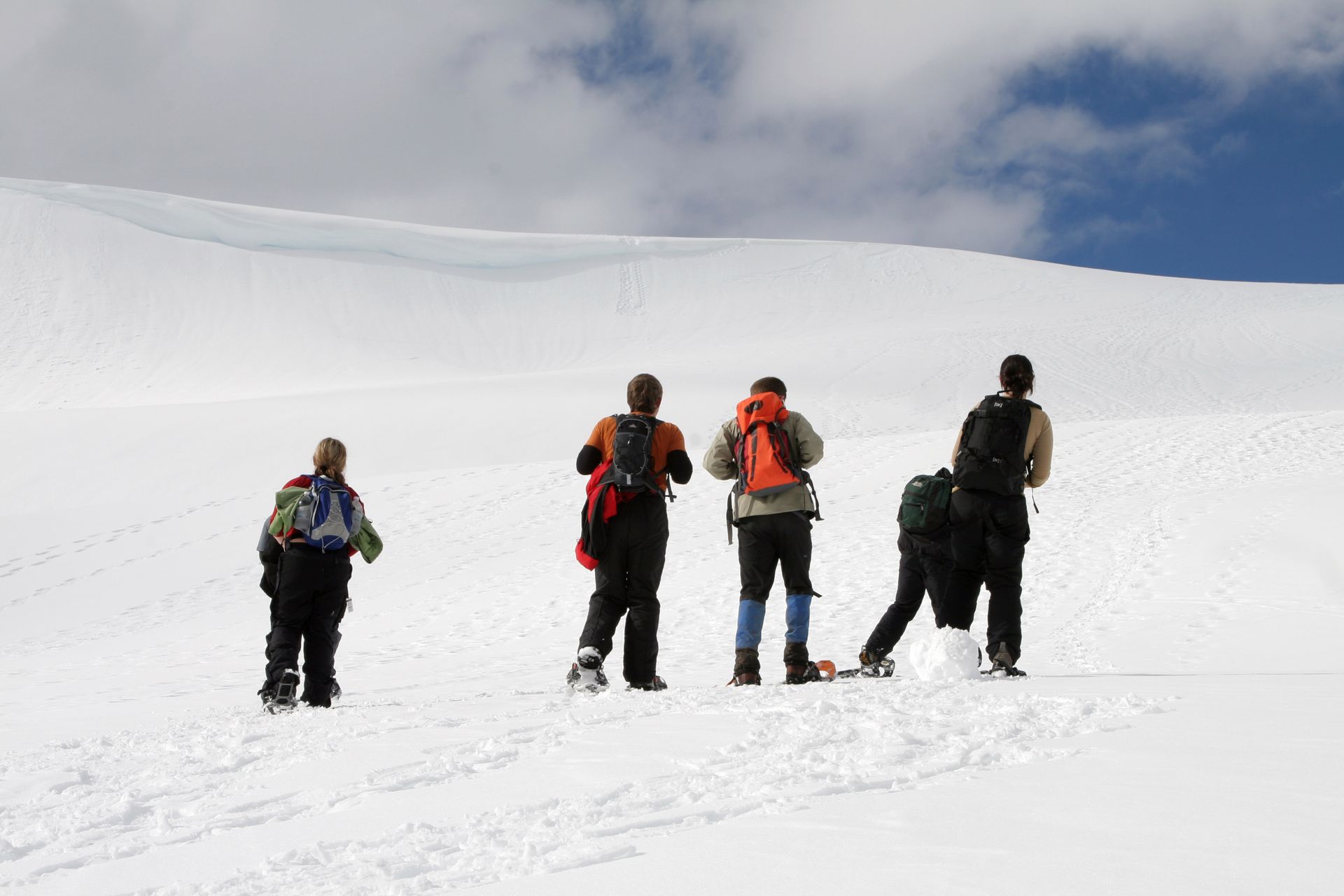 Hikers on a snowy mountain, with backpacks. One is bent over. White snow, blue sky.
