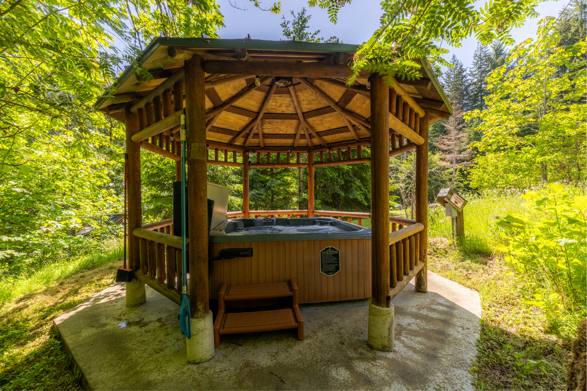 Wooden gazebo with hot tub surrounded by green trees. The gazebo has a brown roof, and the hot tub is dark.
