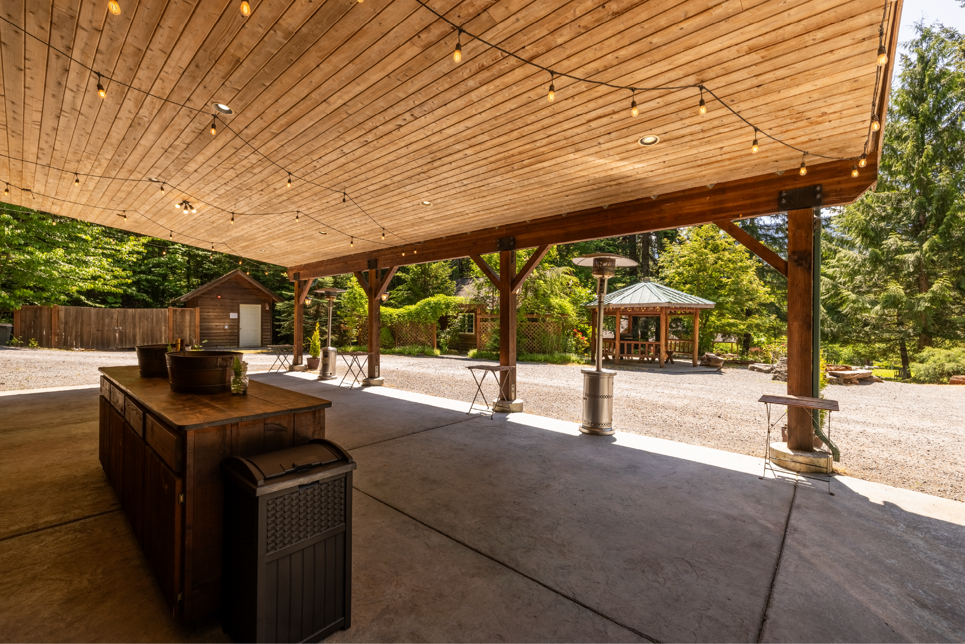 Covered outdoor patio with wooden beams and string lights. A bar, heaters, and gravel ground are visible. A gazebo and trees are in the background.