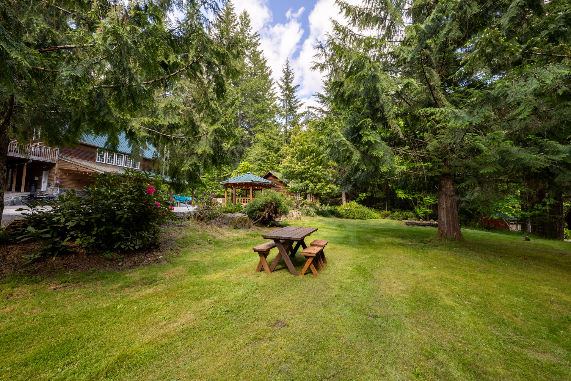 A picnic table sits on a grassy lawn in front of a forest. A gazebo and a building are visible in the background.