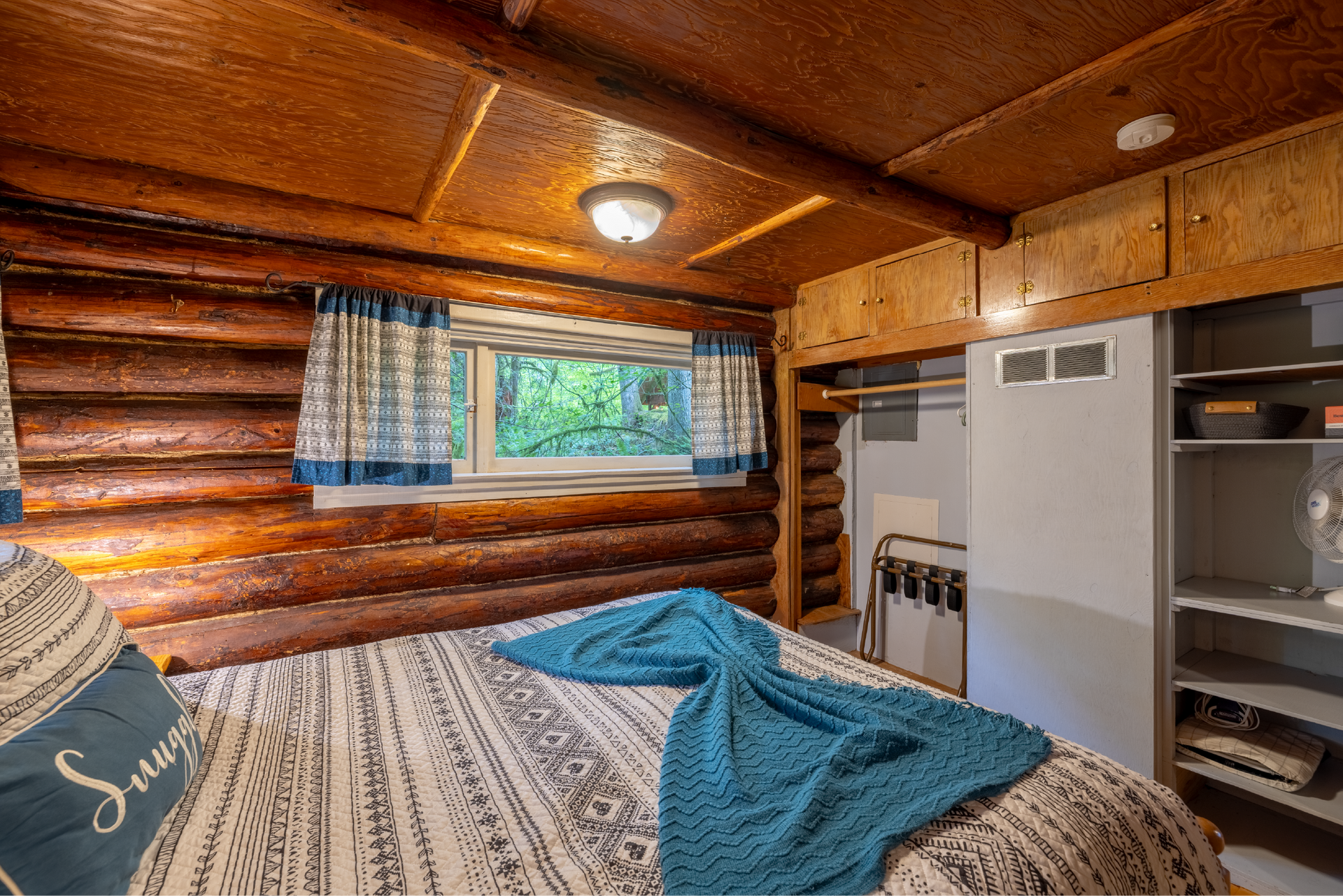 Cozy log cabin bedroom with a bed, closet, and small window overlooking trees. Wooden walls and ceiling, blue accents.