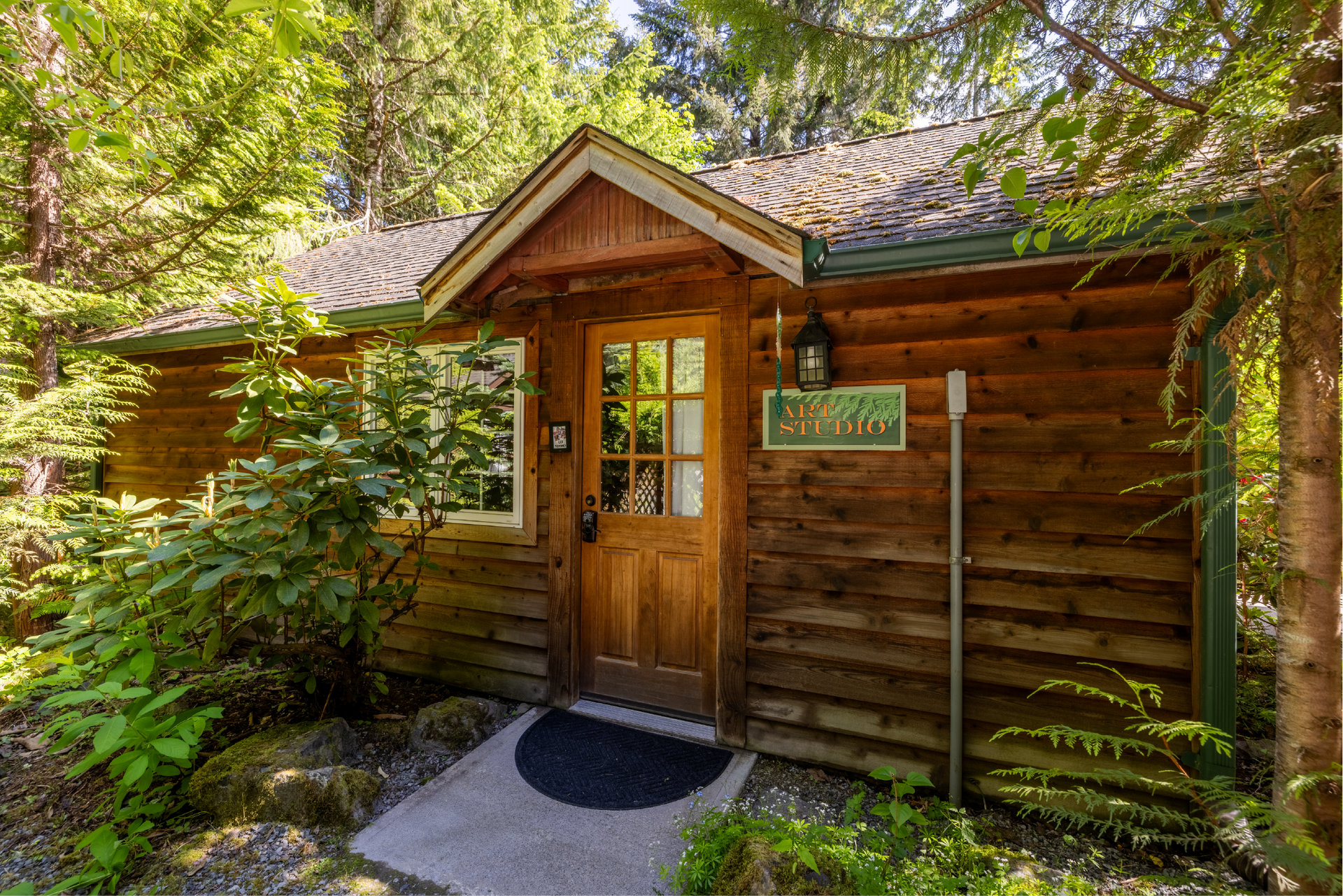 A rustic cabin with a wooden door, surrounded by lush greenery. The building has a sign on the front, and a walkway leading to the door.