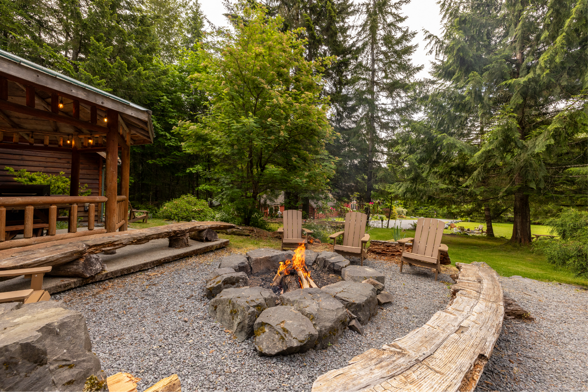 A dog sits near a fire pit surrounded by stone and log seating, next to a log cabin with a picnic table, in a wooded area.