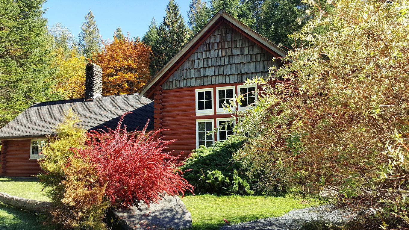 A red log cabin is surrounded by trees and bushes on a sunny day.