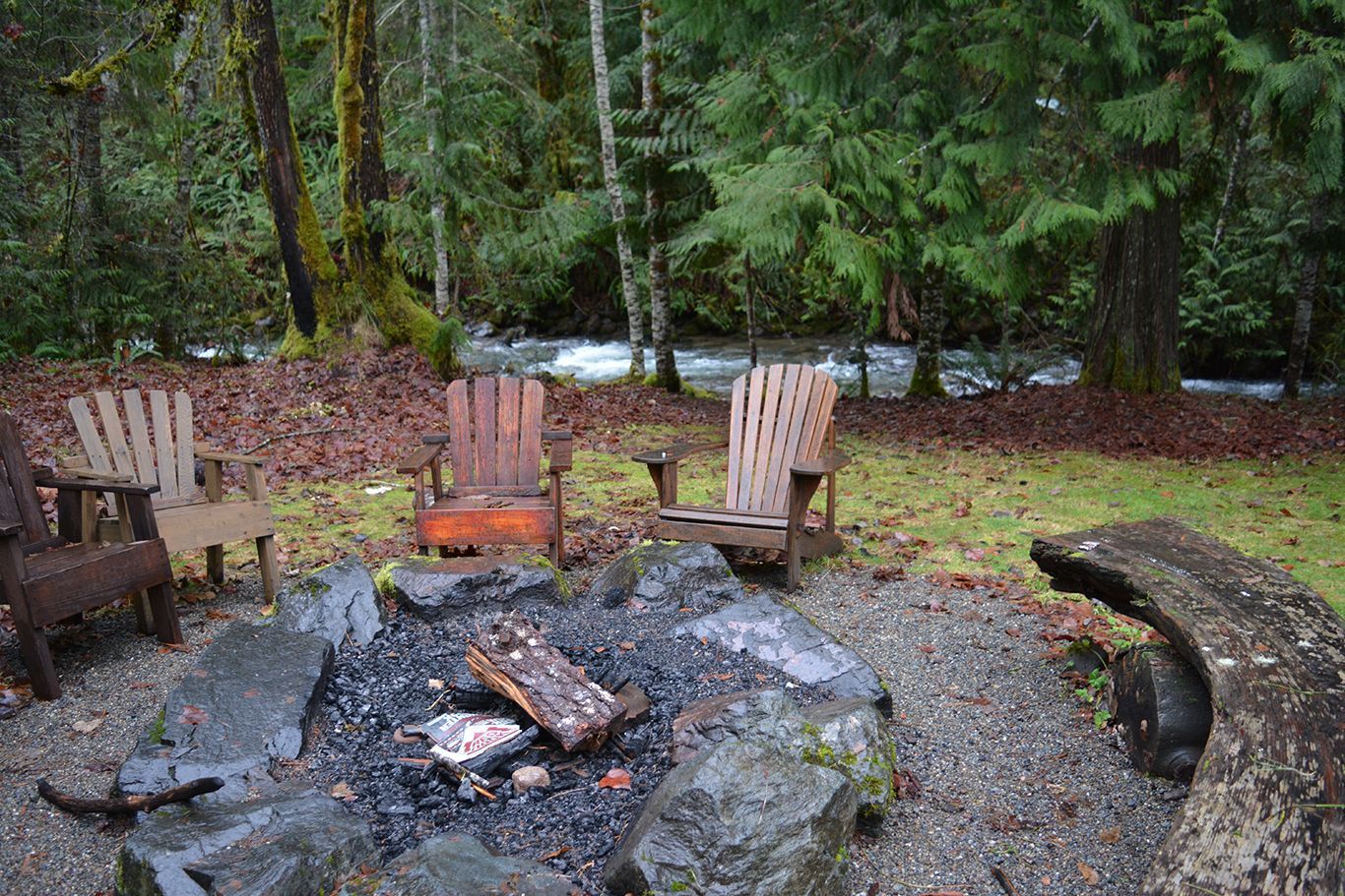 A fire pit in the middle of a forest with chairs around it.
