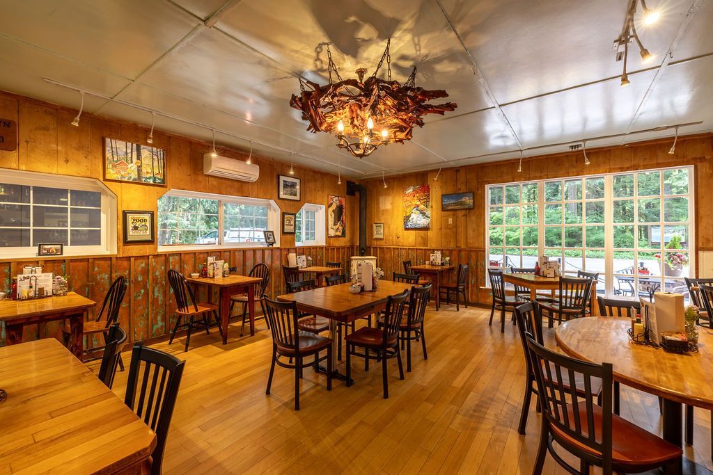 Interior of a rustic restaurant with wooden walls, floors, tables, and chairs. Large windows offer an outdoor view, and a unique chandelier hangs from the ceiling.