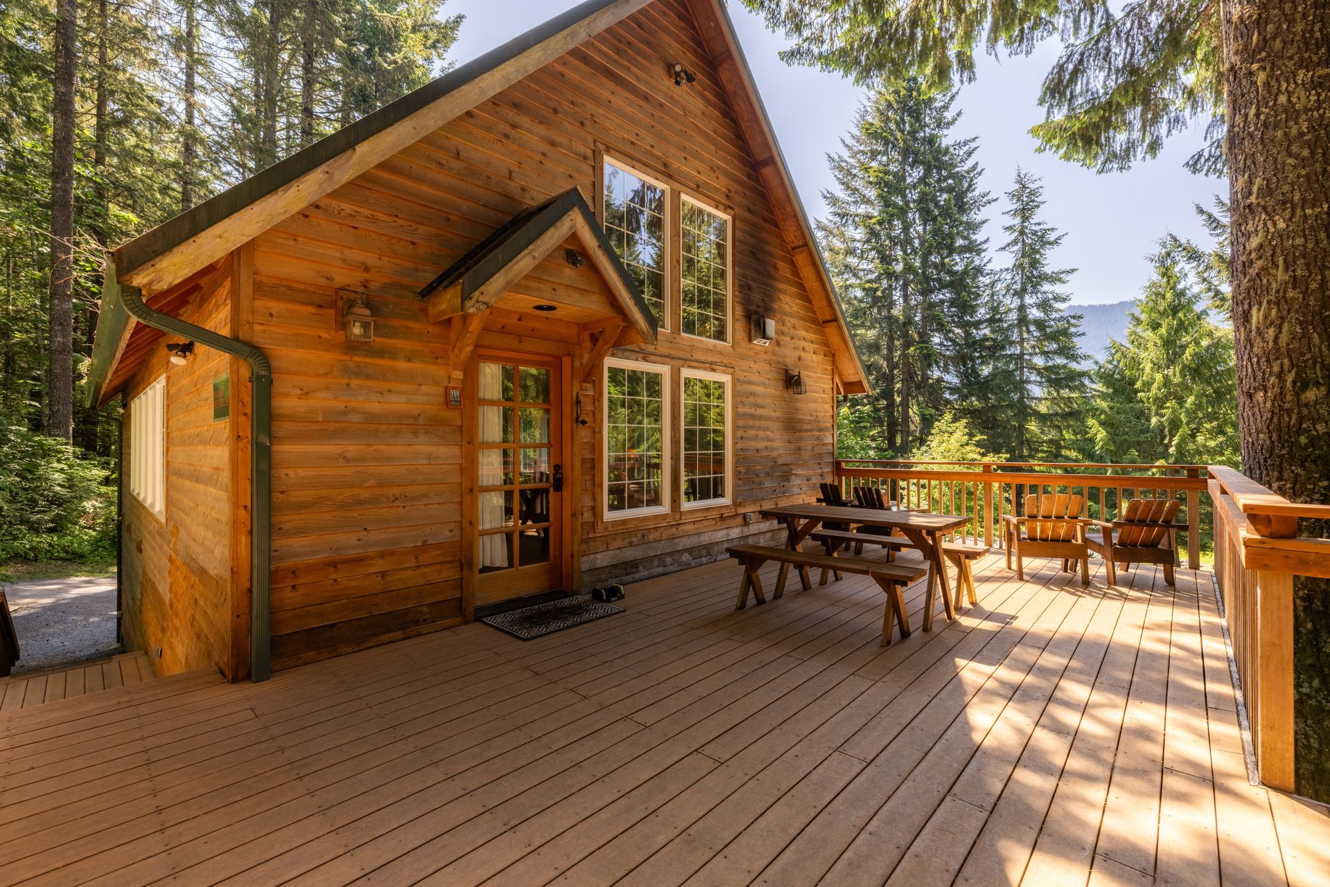Wooden cabin with a deck and outdoor seating in a forest setting. The cabin has large windows and a picnic table.