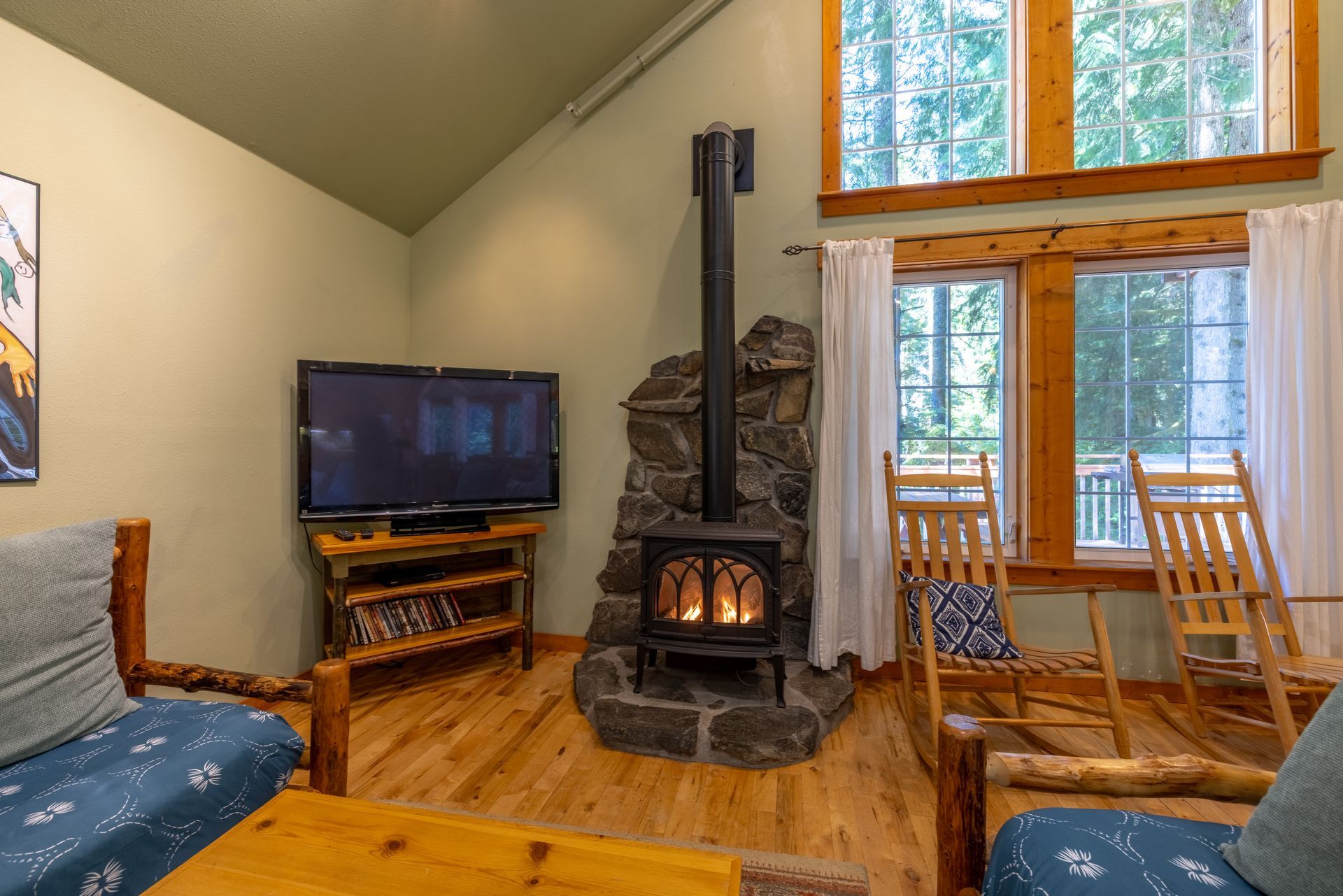 Cozy cabin living room with wood stove, TV, wooden furniture, and large windows with natural light.