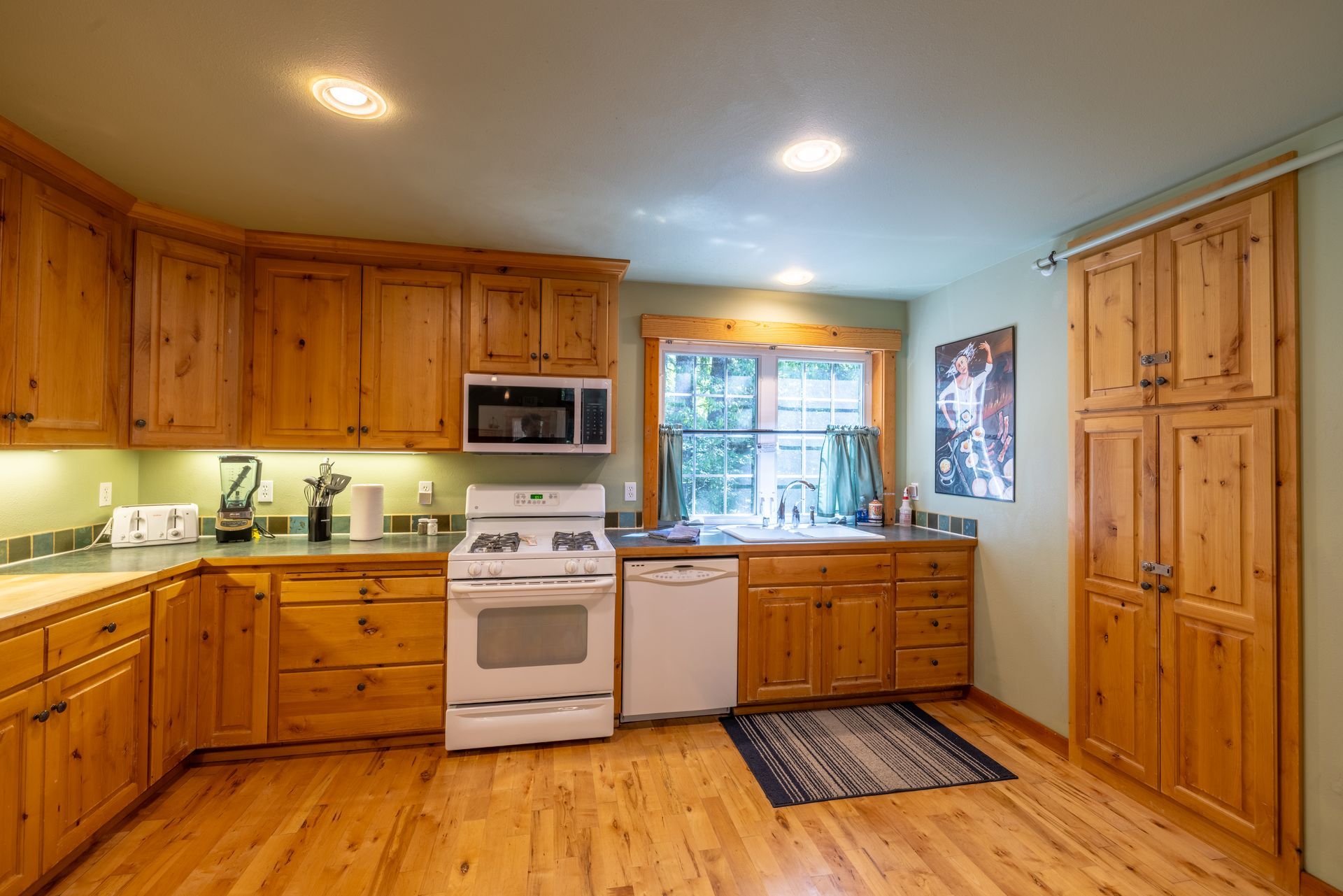 A rustic kitchen with light wood cabinets, a white stove and dishwasher, and wooden floors. The walls are a light green with a small window.