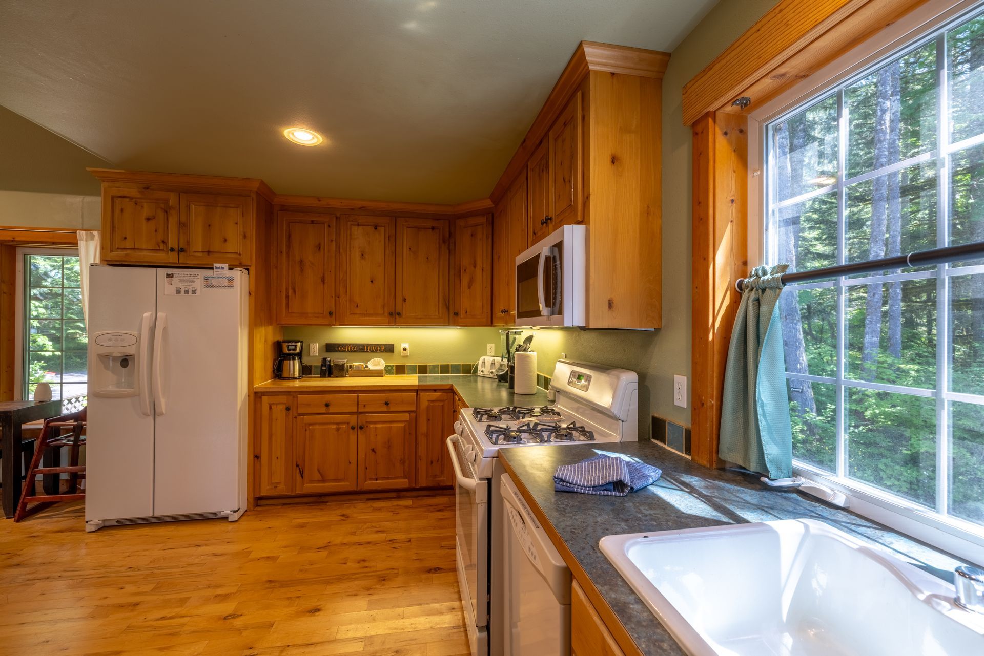 Kitchen with light wood cabinets, white appliances, a sink, and a window overlooking a green outdoors.