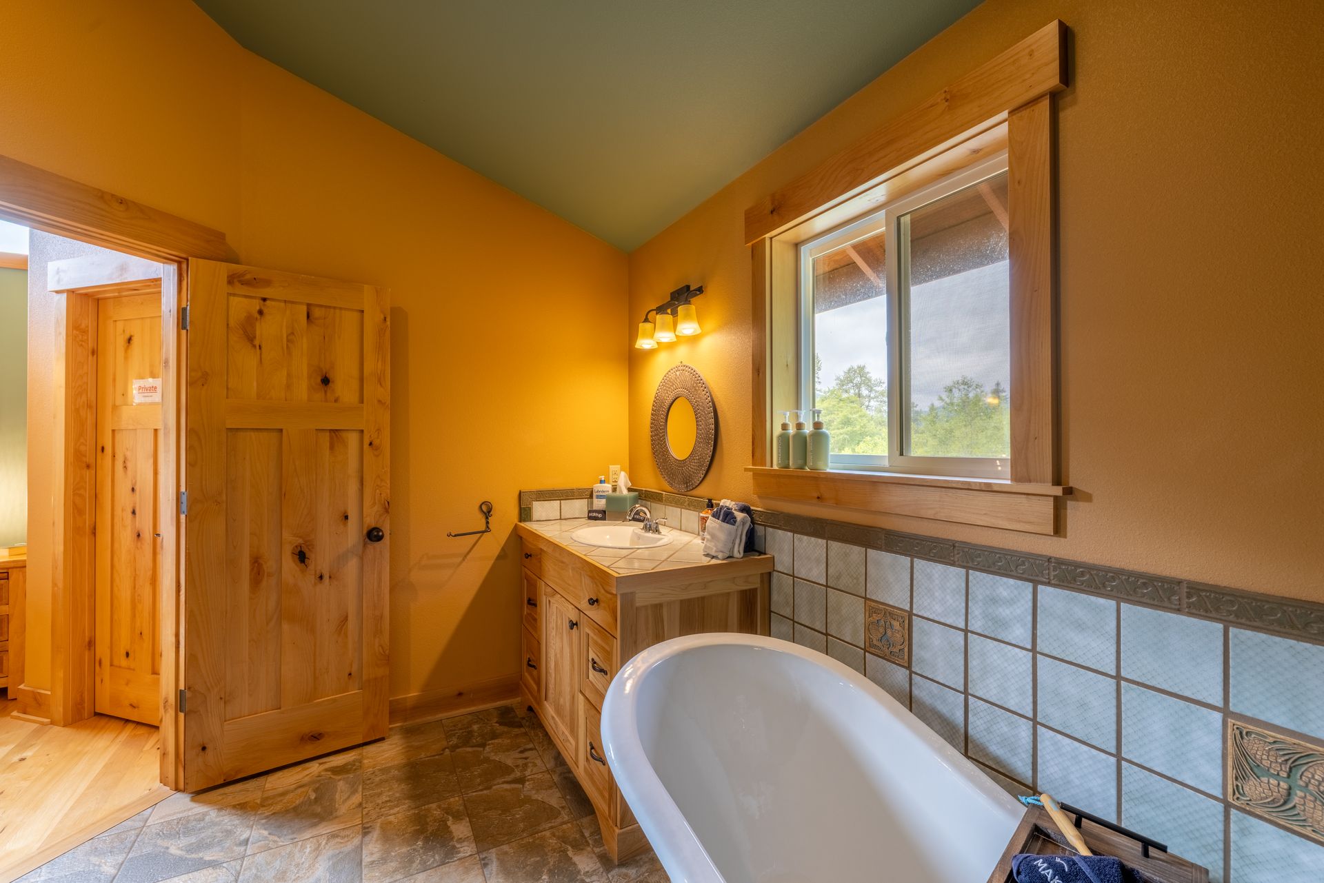 Bathroom with a wooden vanity, white bathtub, and a window. Orange walls, blue ceiling, and wooden trim create a rustic feel.