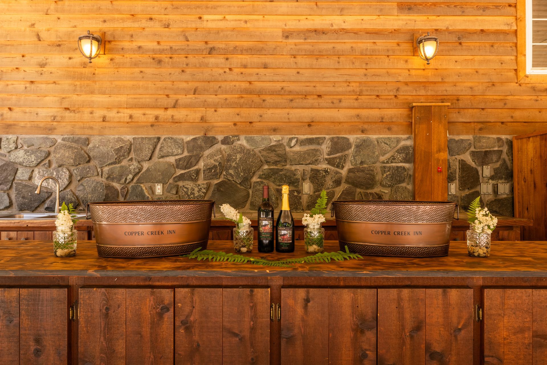 A bar counter with two copper ice buckets, wine bottles, and floral arrangements against a wooden and stone backdrop.