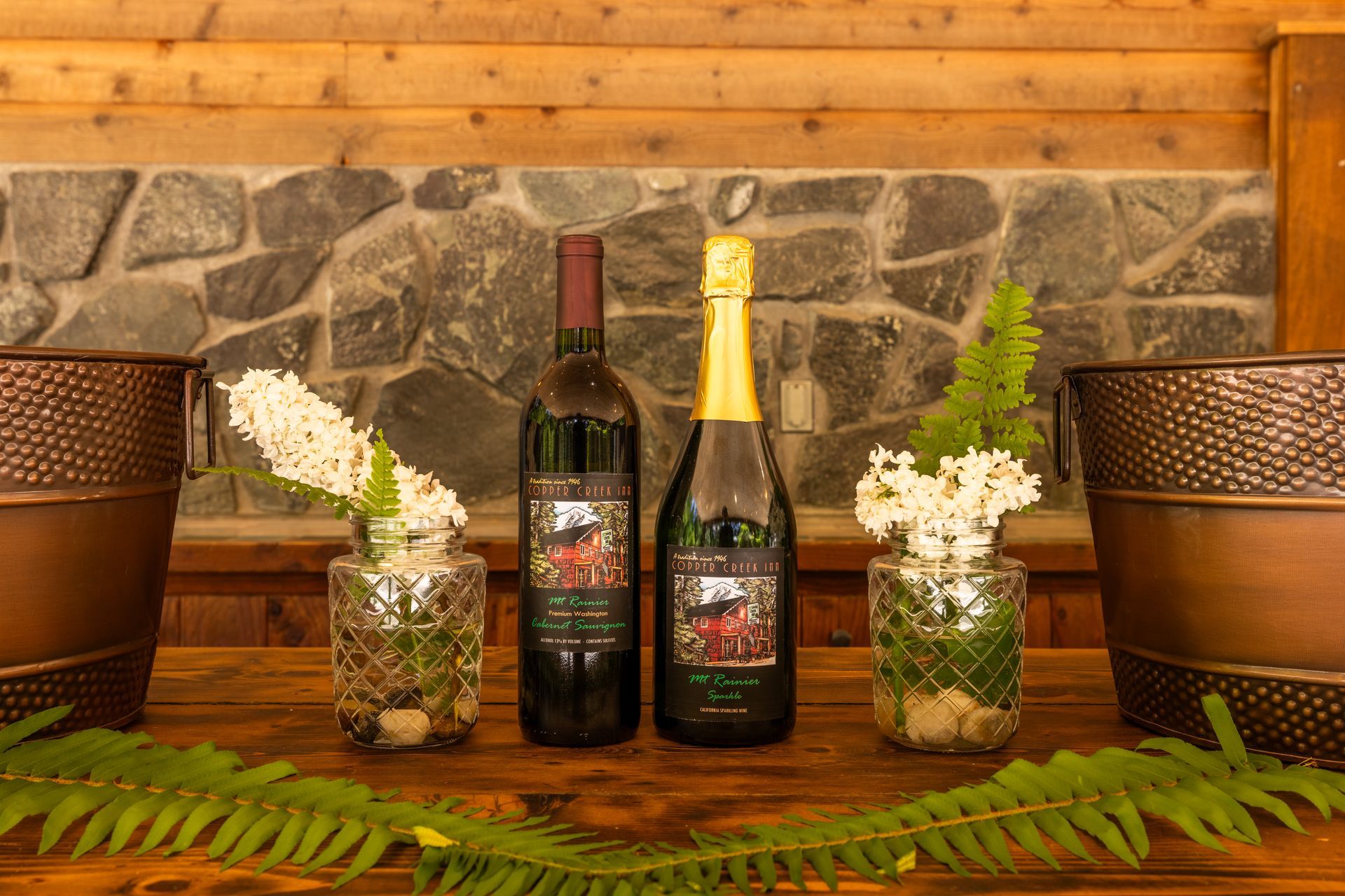 Two wine bottles, red and champagne, with flower vases and ferns on a wooden table in front of a stone wall.