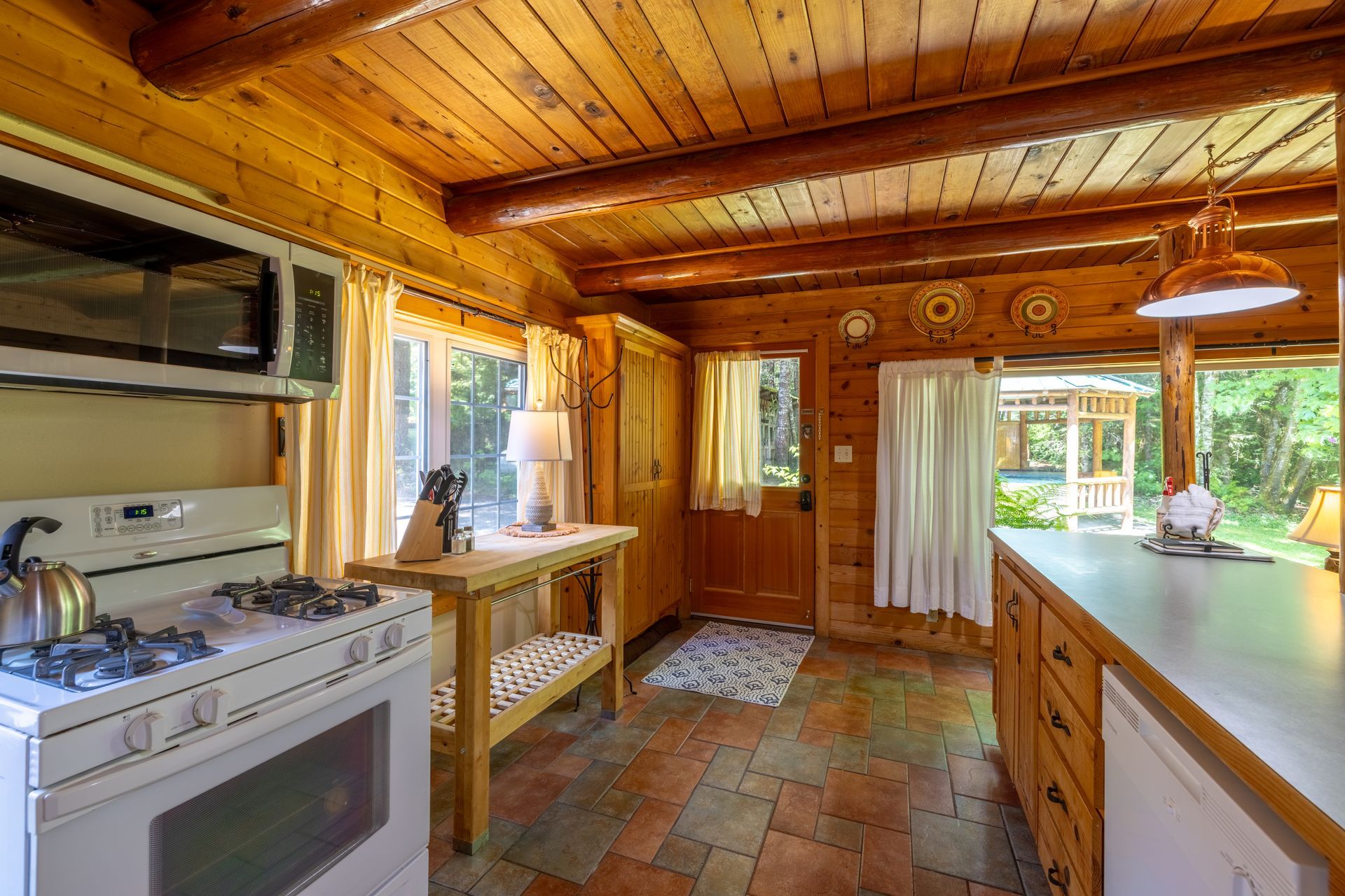 Cozy cabin kitchen with wood paneling, brick floor, and appliances. A gazebo is visible through a doorway.