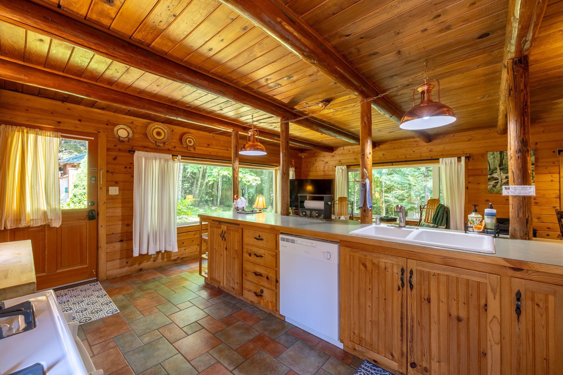 Rustic kitchen with wooden walls and cabinets. White appliances and two pendant lights are visible. Windows provide a view of greenery.